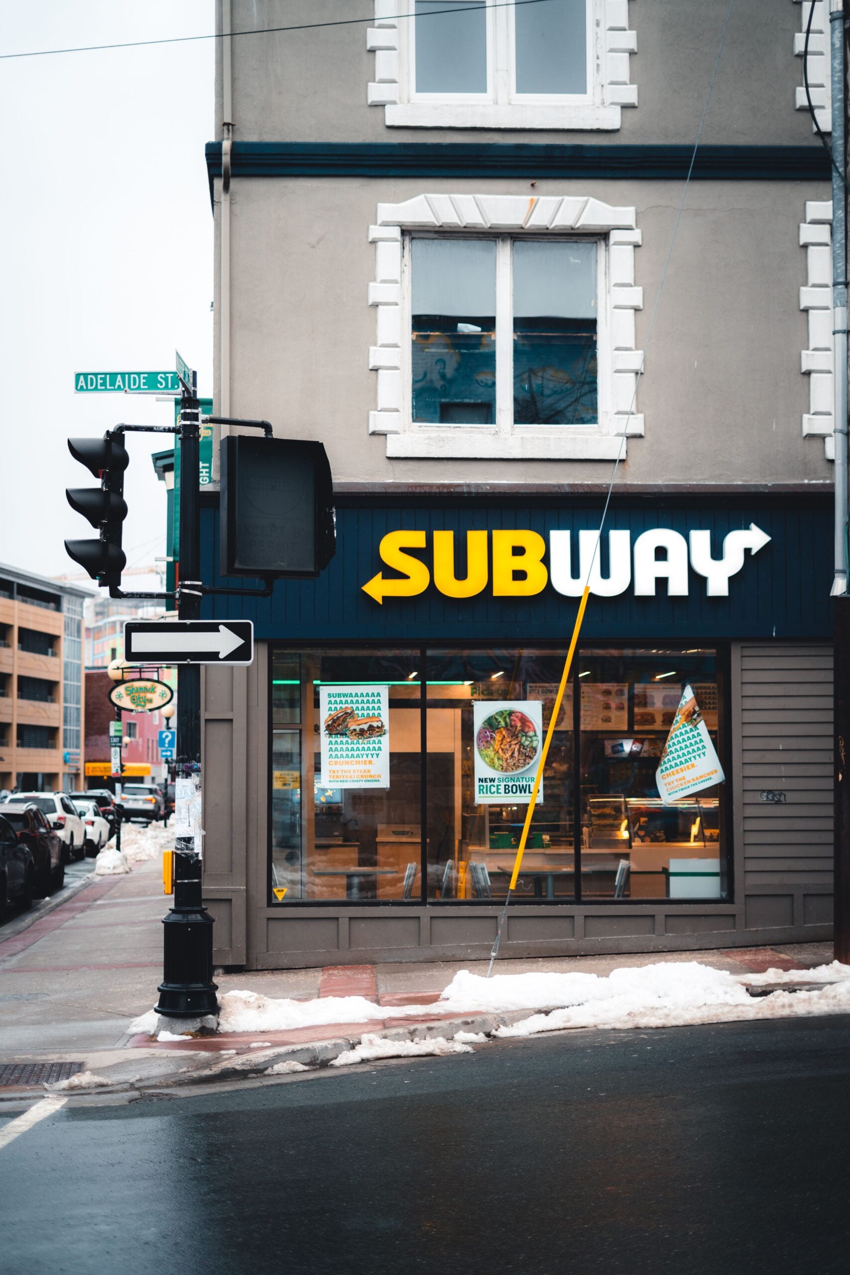 A Subway restaurant on a city corner with snow on the ground and a street sign reading “Adelaide St.” Large Subway logo and food posters are visible on the window.