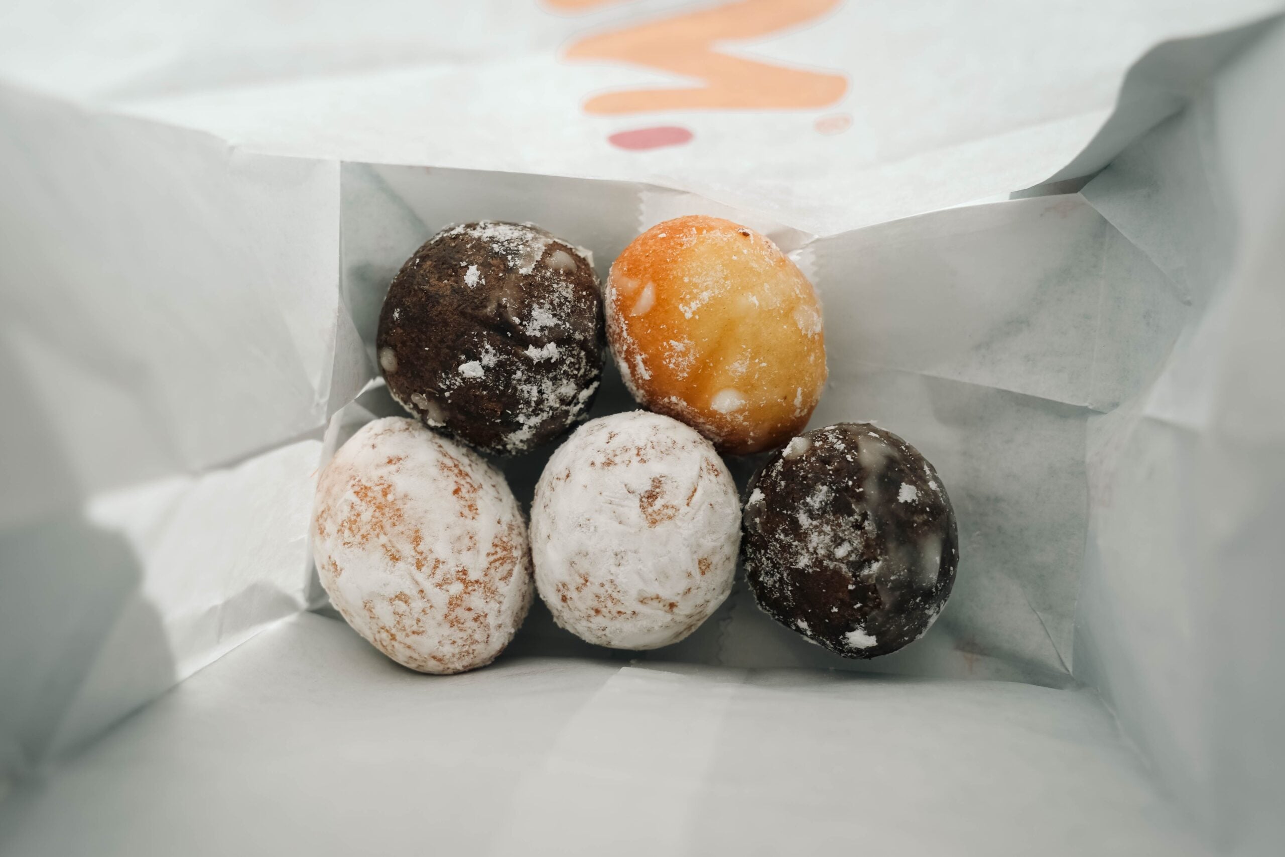 Five powdered donut holes, some chocolate and some plain, sit inside a white paper bag, viewed from above.