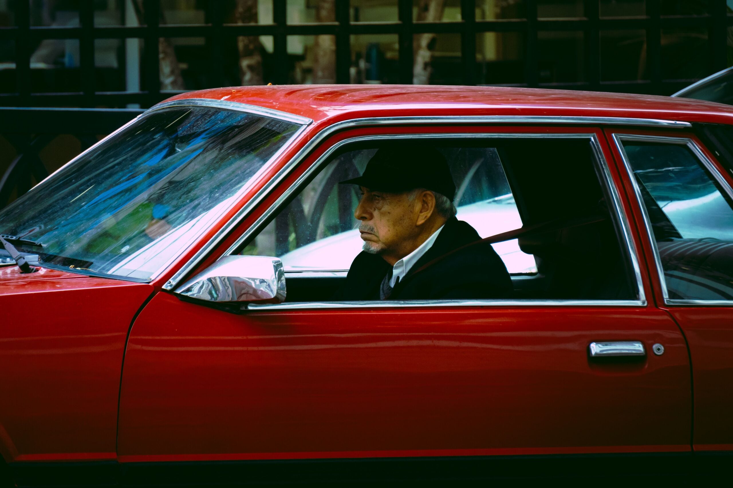 An older man wearing a cap and suit sits in the driver’s seat of a red vintage car, looking straight ahead with a serious expression—a fitting image of how boomers saving money appreciate timeless values and classic style.