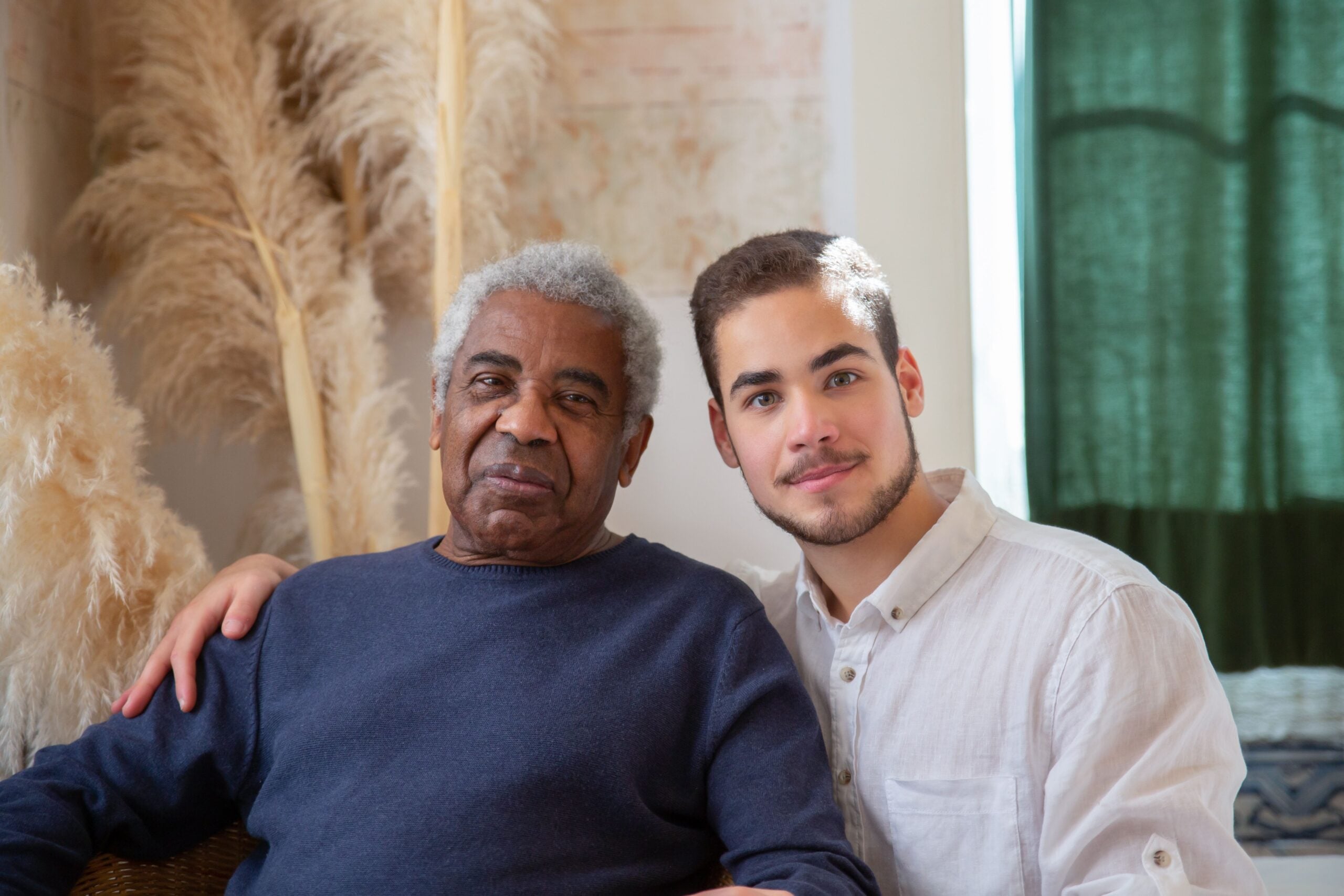 An older man and a younger man sit close together indoors, both smiling gently at the camera. The younger man has his arm around the older man as they share a warm moment. Pampas grass decorates the background, adding a modern touch to their setting—perhaps making it the perfect spot to exchange some Boomer facts between generations.