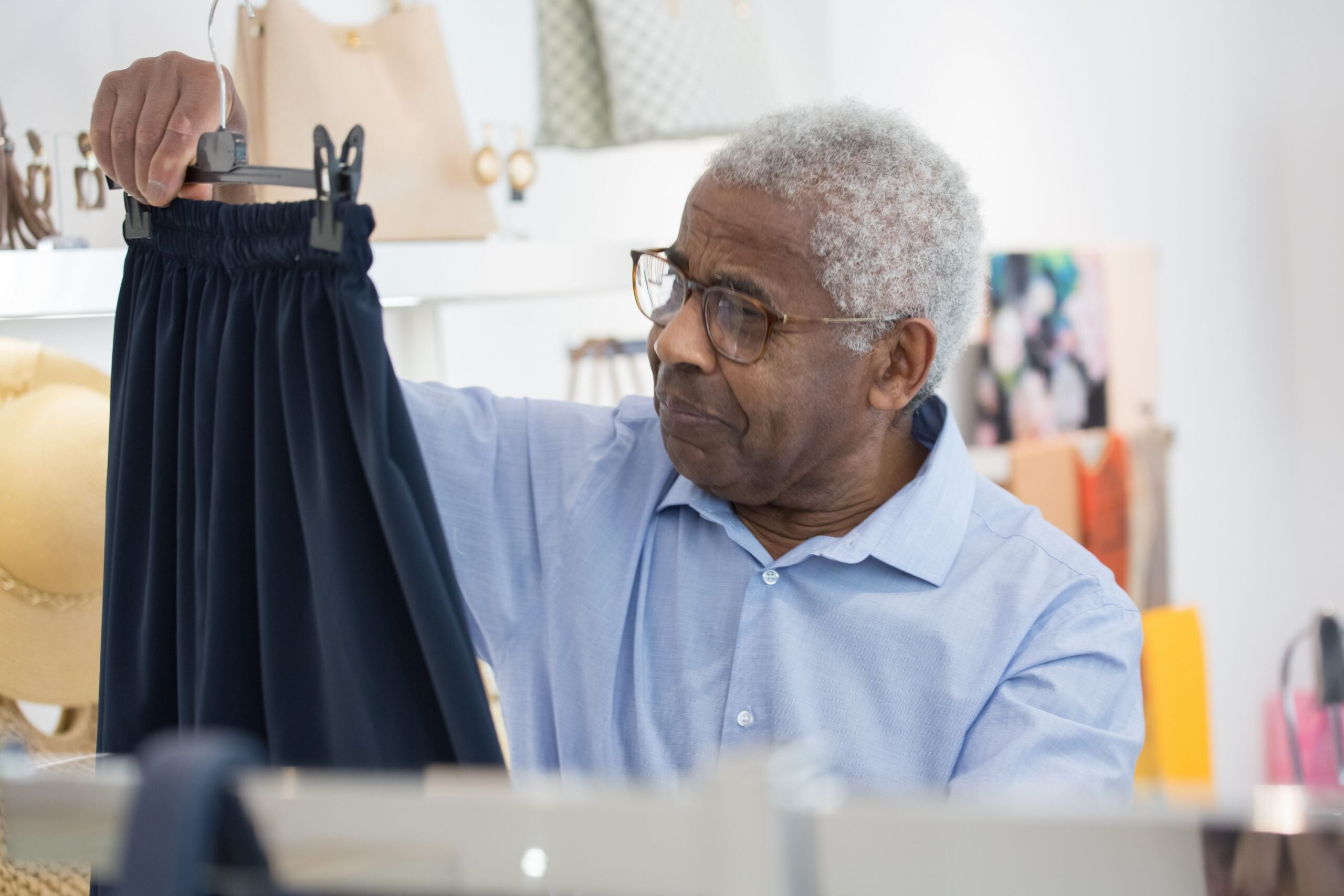 An older man in glasses and a light blue shirt examines a navy blue skirt on a hanger in a clothing store, embodying how boomers saving money carefully consider their purchases before buying.