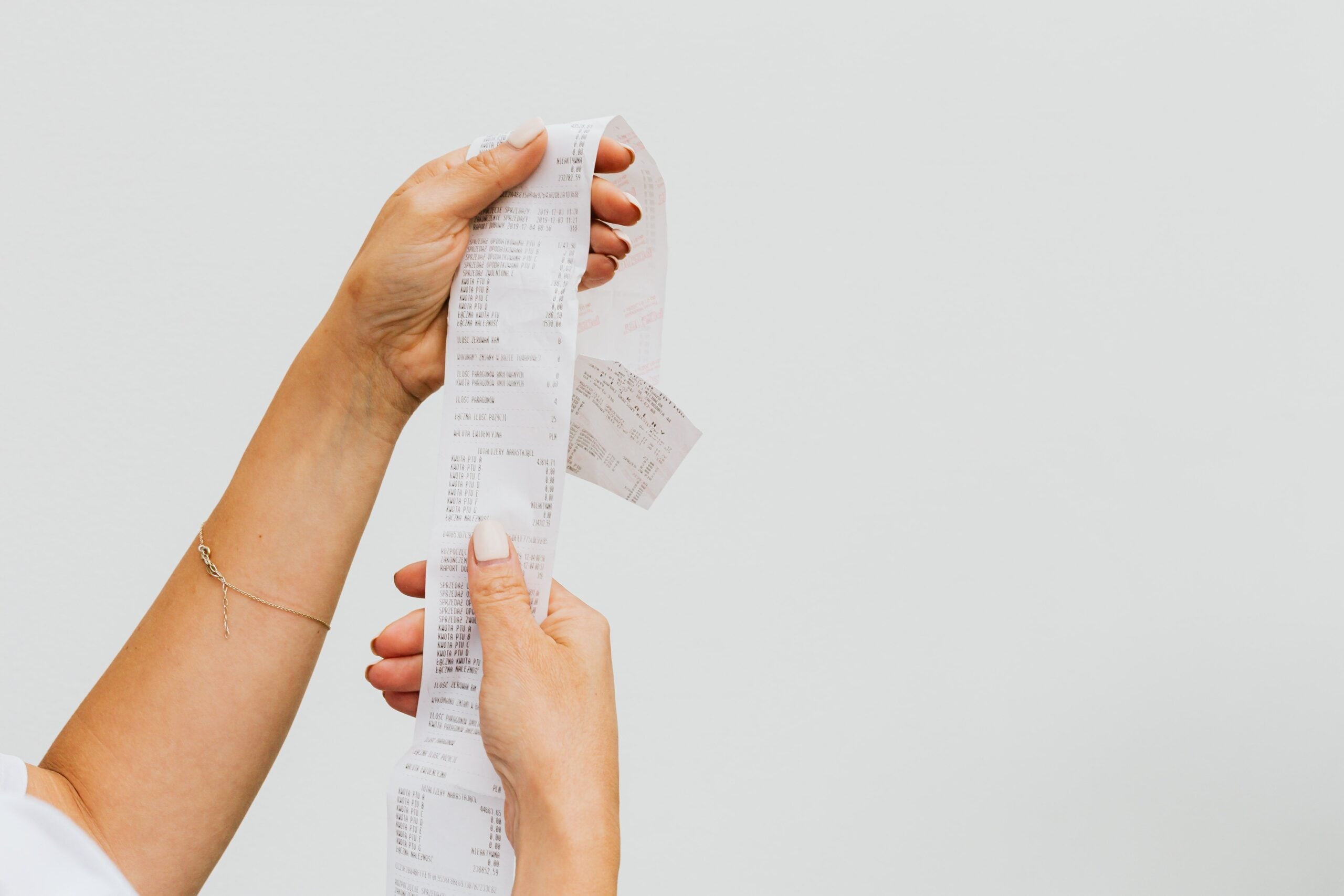 A person holding and inspecting a long printed receipt with both hands against a plain white background, carefully checking for any travel destination red flags among the listed expenses.
