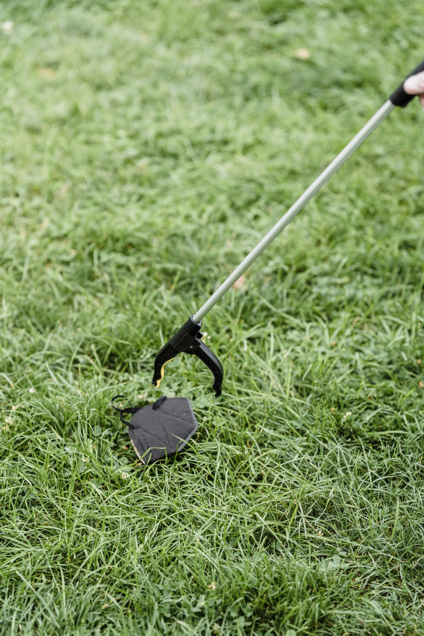 A person uses a litter picker grabber to pick up a black face mask from green grass outdoors.