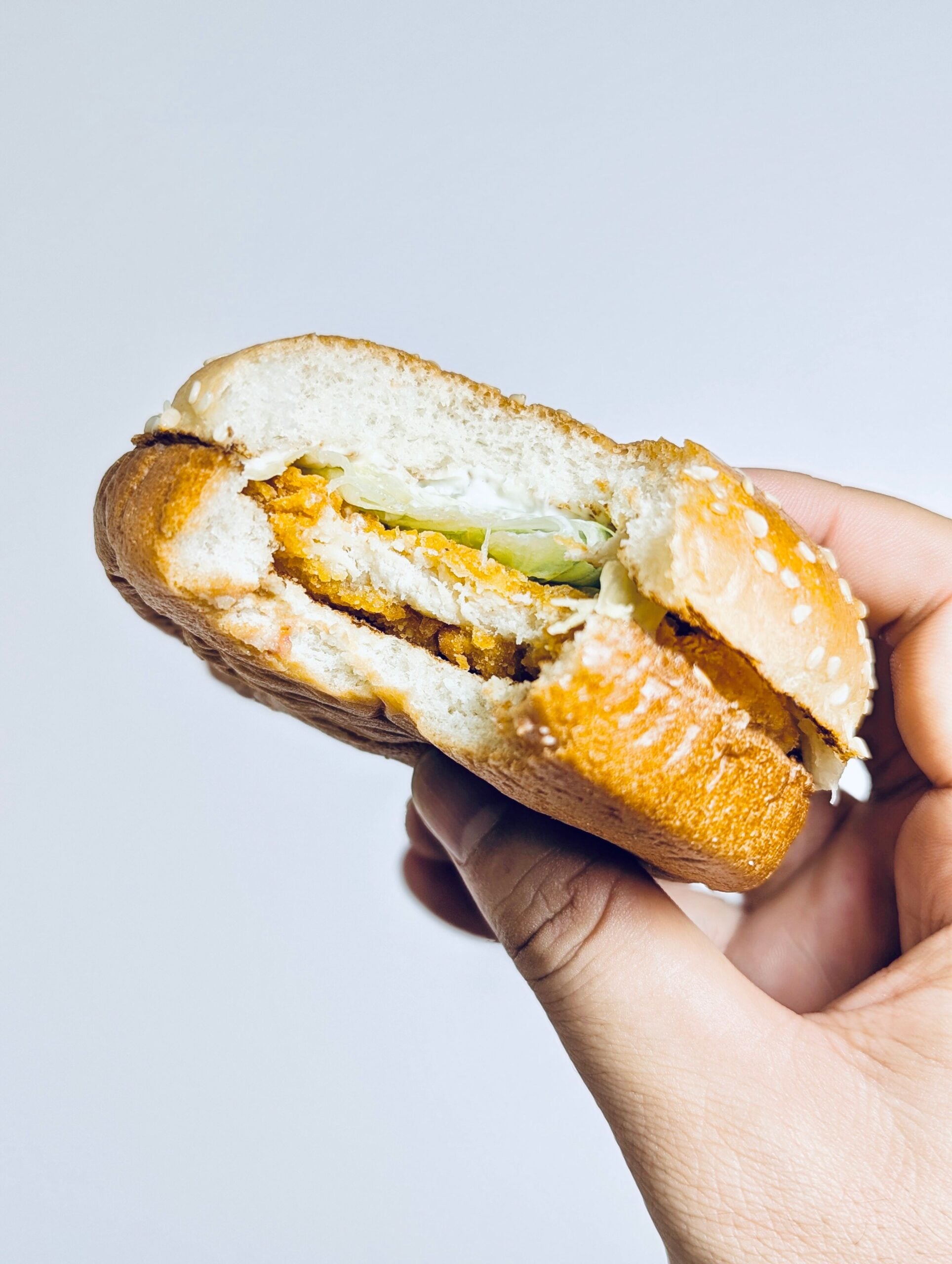 A hand holding a bitten chicken sandwich with lettuce and sauce on a sesame seed bun against a plain white background.