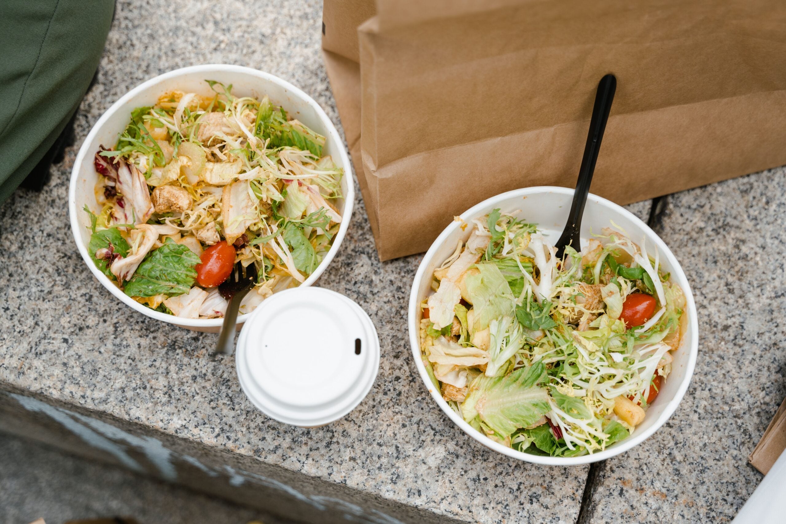 Two bowls of mixed salad with lettuce, tomatoes, and shredded chicken sit on a stone surface next to a paper coffee cup and a brown paper bag.