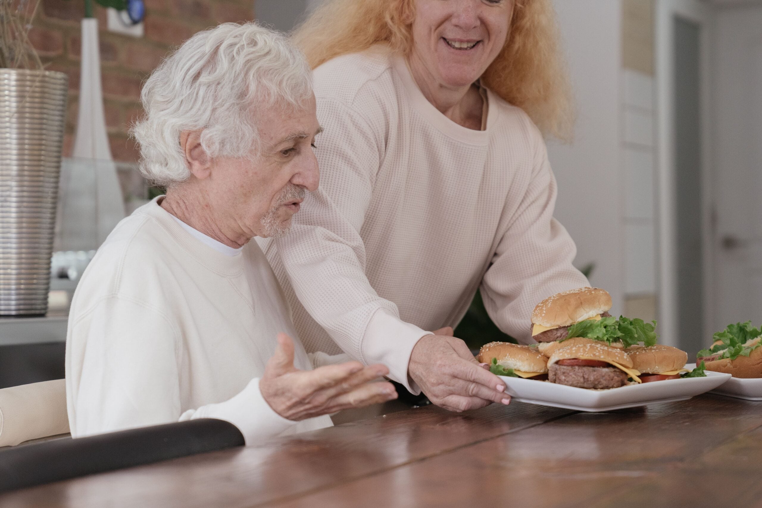 An older man sits at a table while a woman offers him a plate of hamburgers, both smiling. Indoors, the scene looks friendly and inviting—a perfect example of boomers saving money by enjoying homemade meals together.