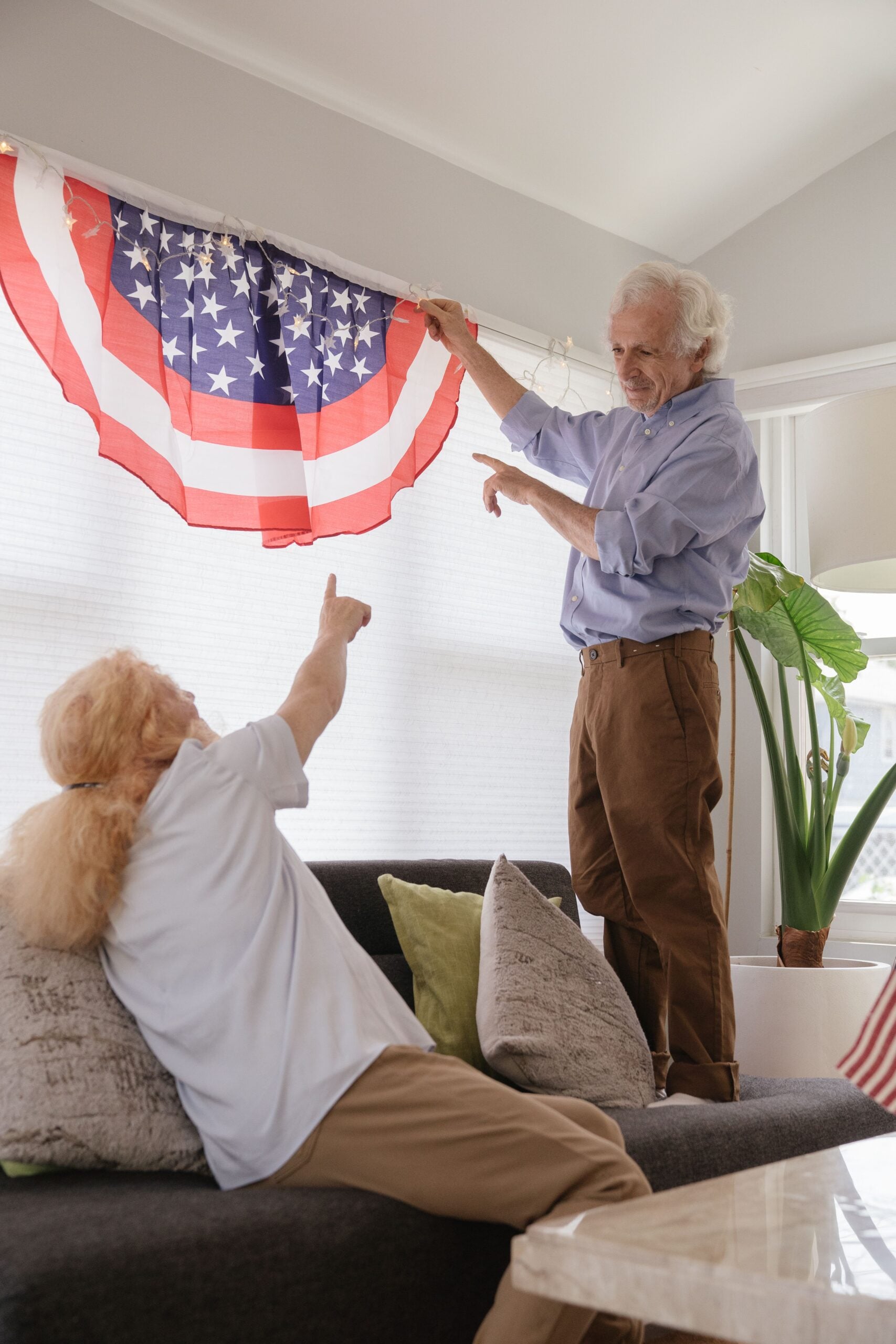 An older man stands on a couch, hanging a red, white, and blue patriotic decoration, while an older woman points and smiles at him—two boomers saving money by decorating their home themselves.