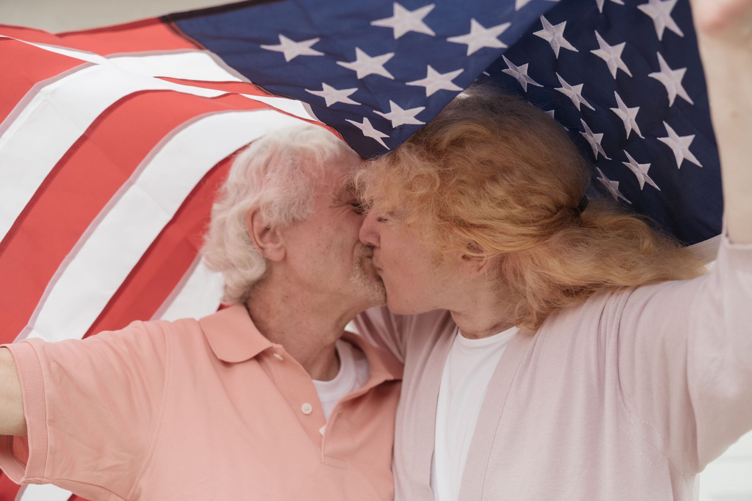 Two older adults share a joyful kiss while holding an American flag above their heads, partially covering them, in a scene that radiates happiness and intimacy. This heartwarming moment captures not just affection, but also celebrates boomer facts—reminding us that love and pride endure through every generation. The bright background further emphasizes the couple’s lively connection and patriotic spirit.