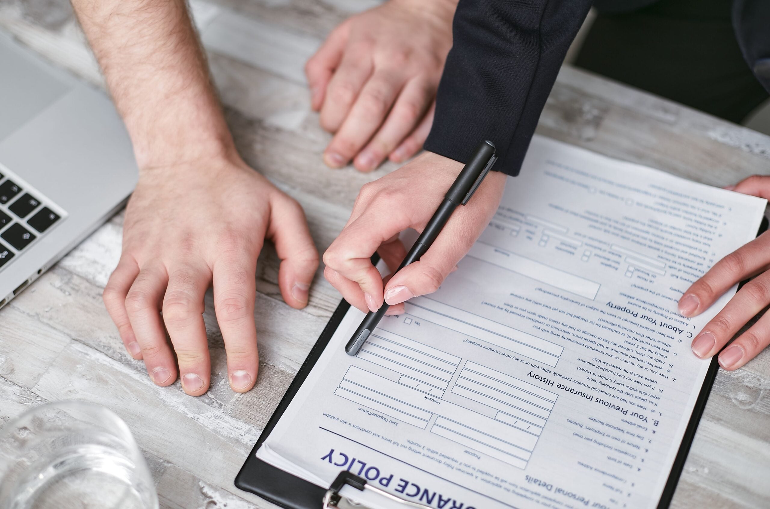 Two people review and fill out an insurance policy form on a clipboard at a desk, with one holding a pen and the other placing a hand nearby next to a laptop, carefully considering any travel destination red flags before finalizing their coverage.