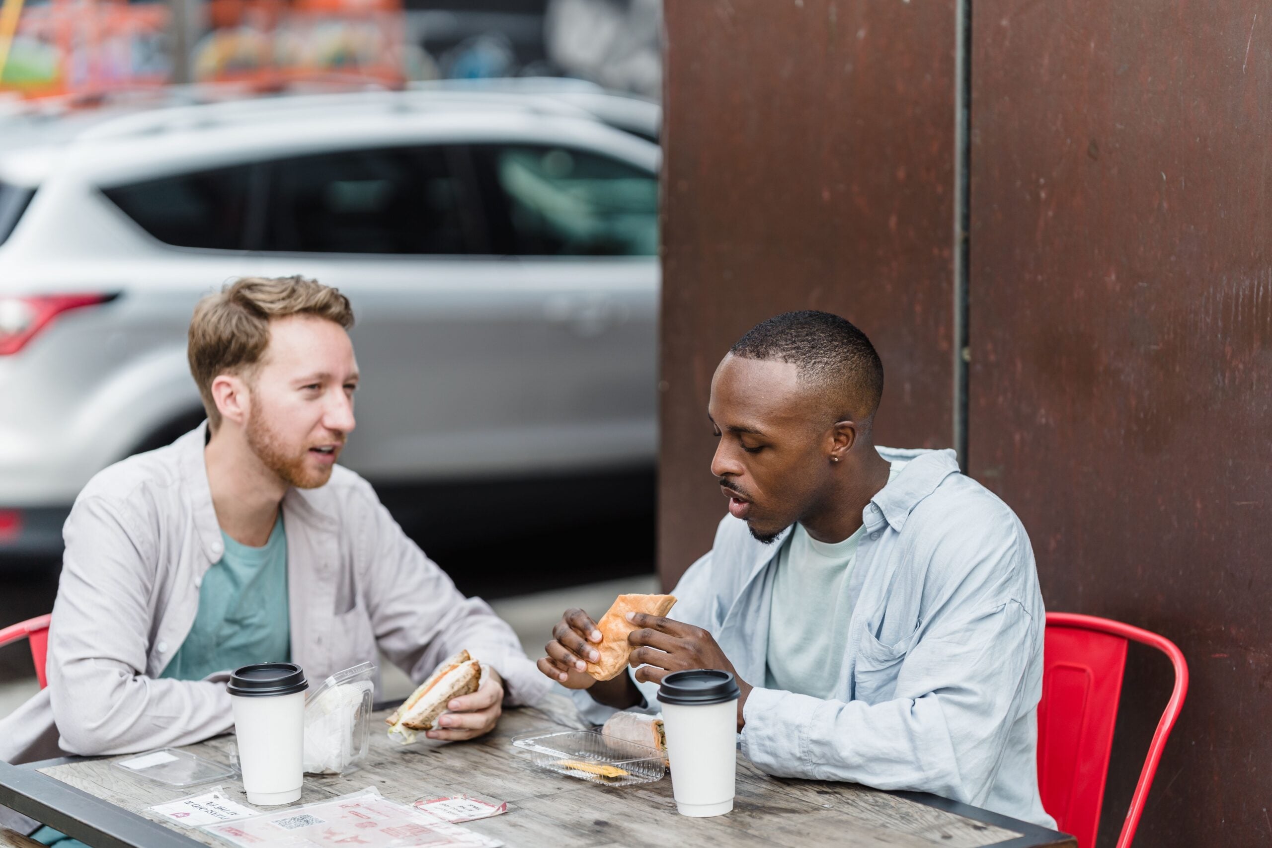 Two men sit at an outdoor table, drinking coffee and eating sandwiches. They are engaged in conversation. A parked car and an urban background are visible behind them.