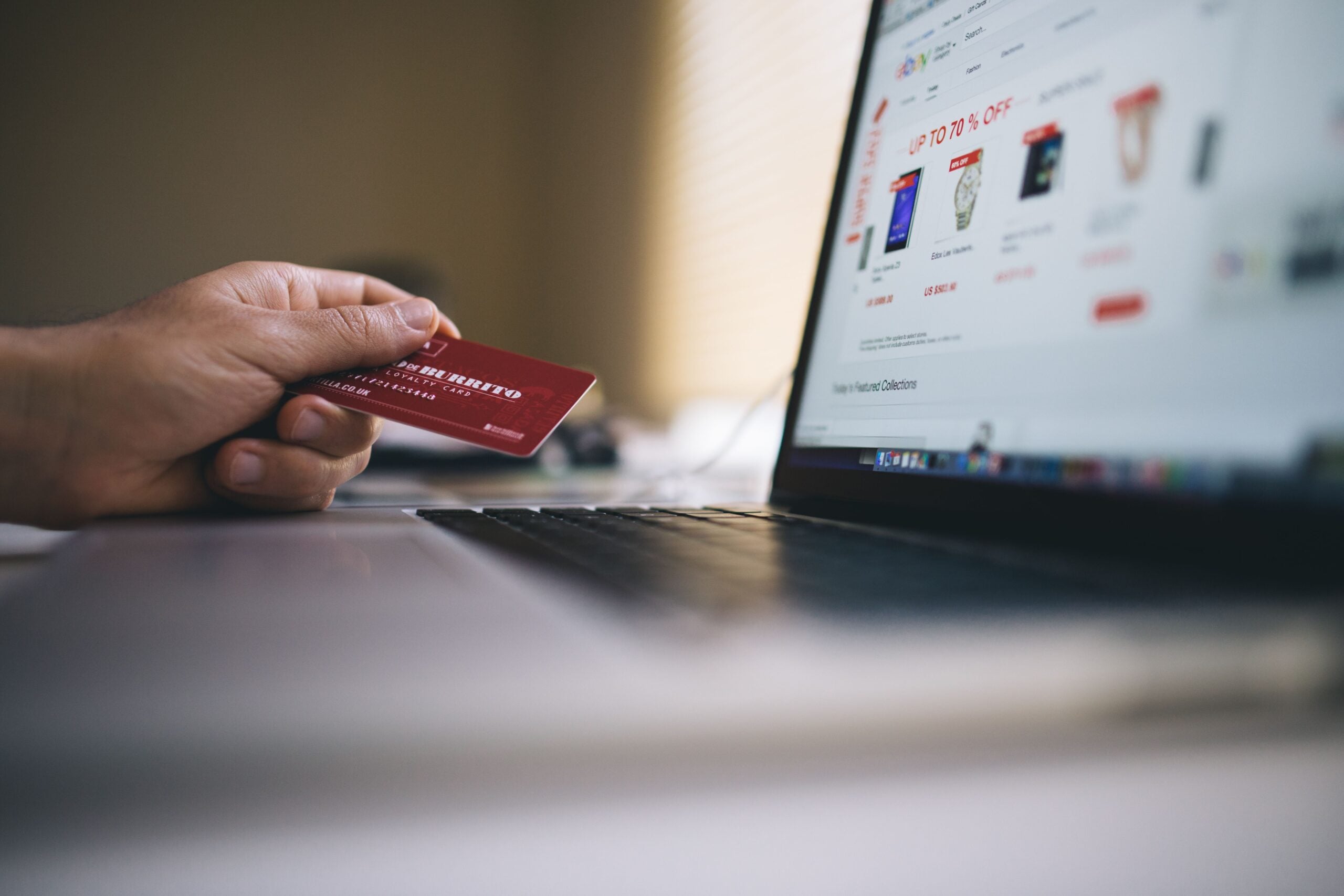 A person holds a credit card in one hand while using a laptop to shop online. The laptop screen displays a shopping website with product images, discounts, and even highlights travel destination red flags to help shoppers make informed choices.
