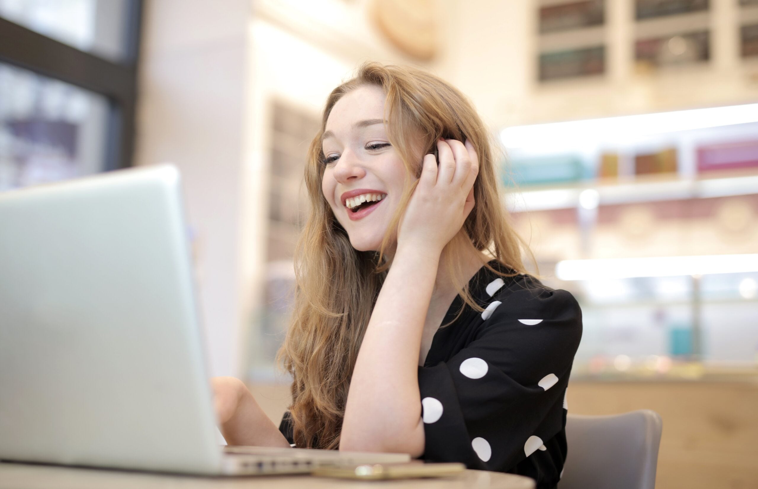 Smiling woman with long hair in a black polka dot shirt sits at a table, looking at a laptop screen, appearing happy and engaged in a bright, modern indoor setting.