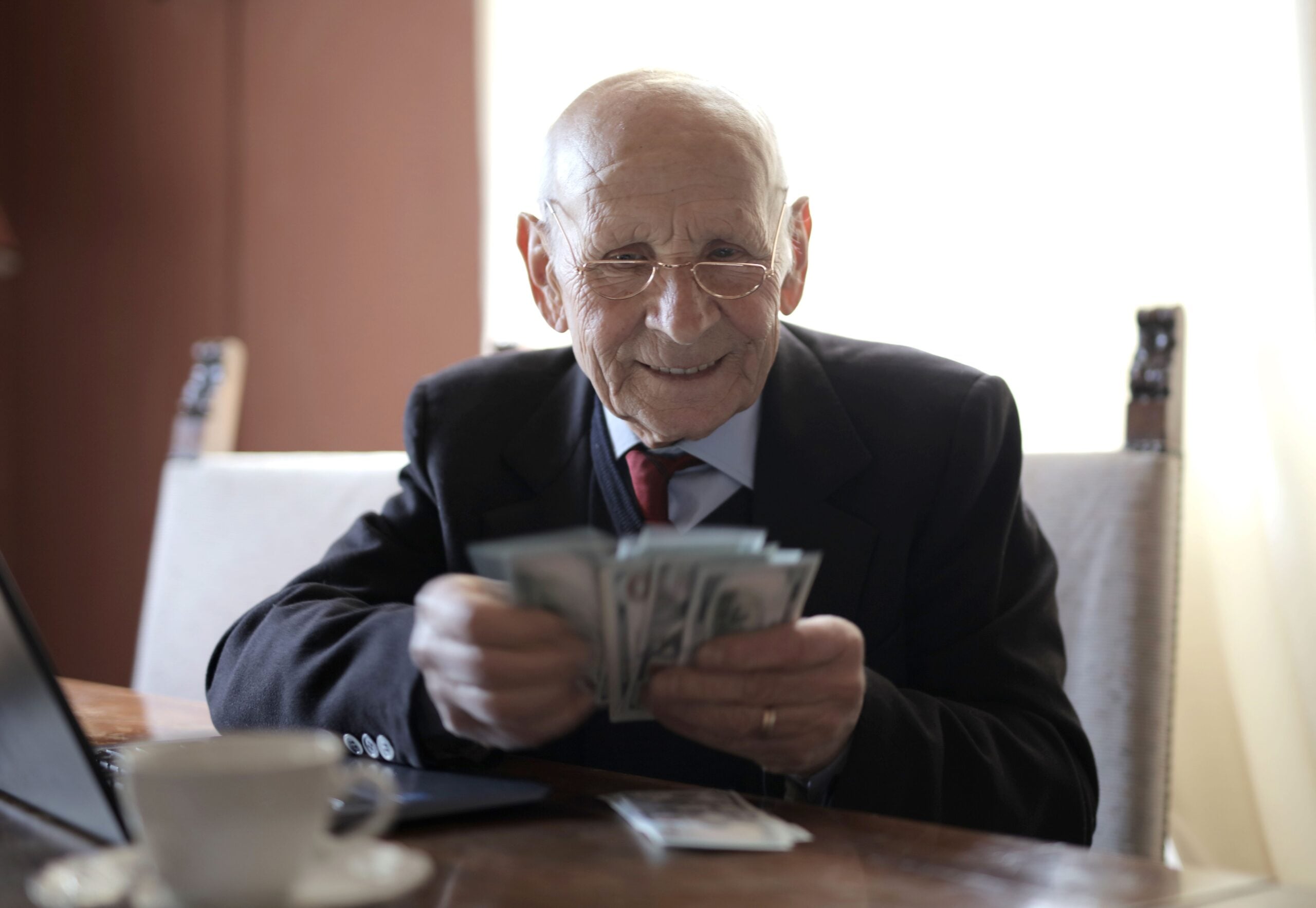 An elderly man in a suit and glasses sits at a table, smiling while holding and counting a stack of dollar bills, with a laptop nearby—an image that brings to mind classic Boomer facts about financial savvy and lifelong saving habits.