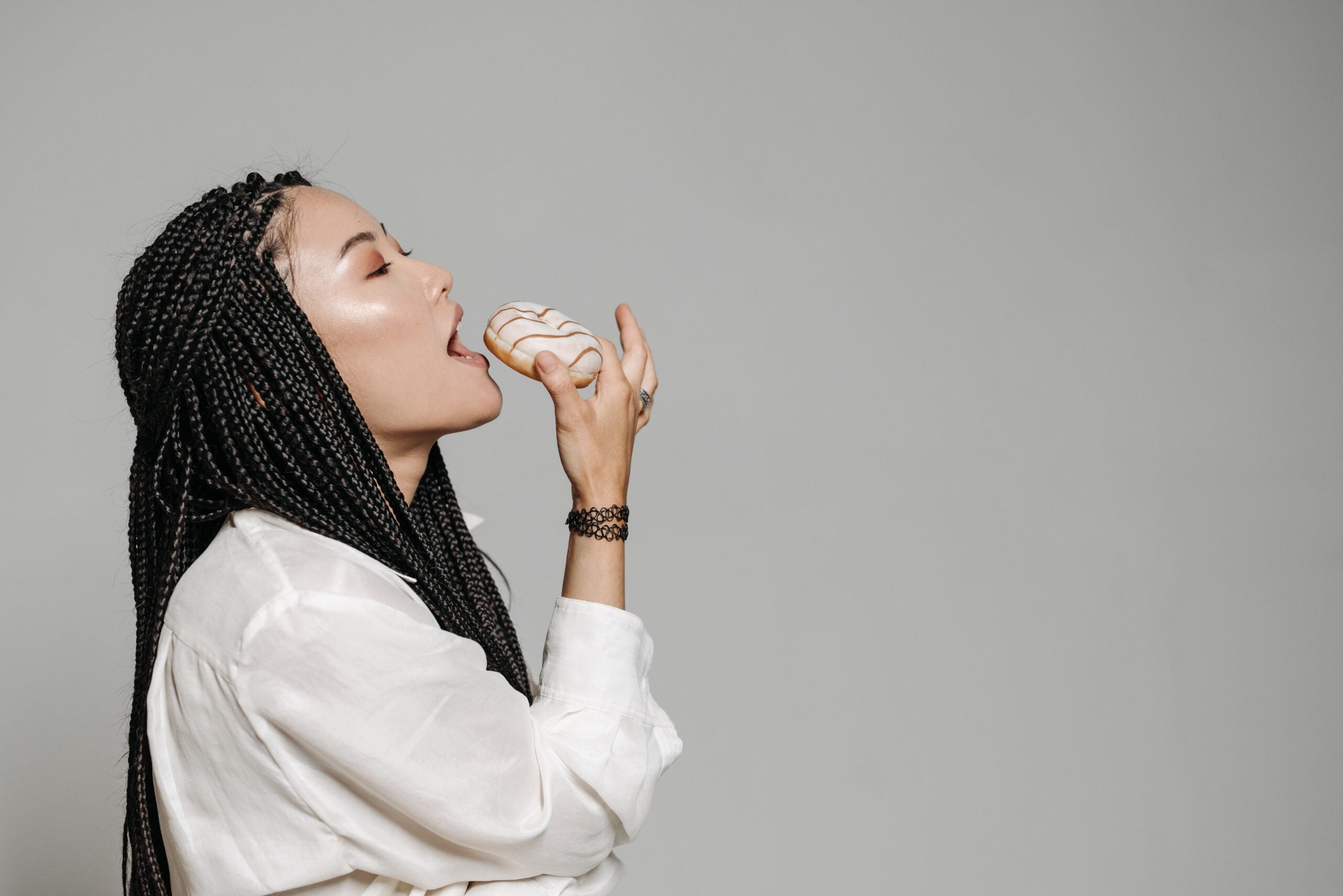 Woman with braids in a white shirt is holding a decorated donut near her mouth, appearing about to take a bite, against a plain gray background.