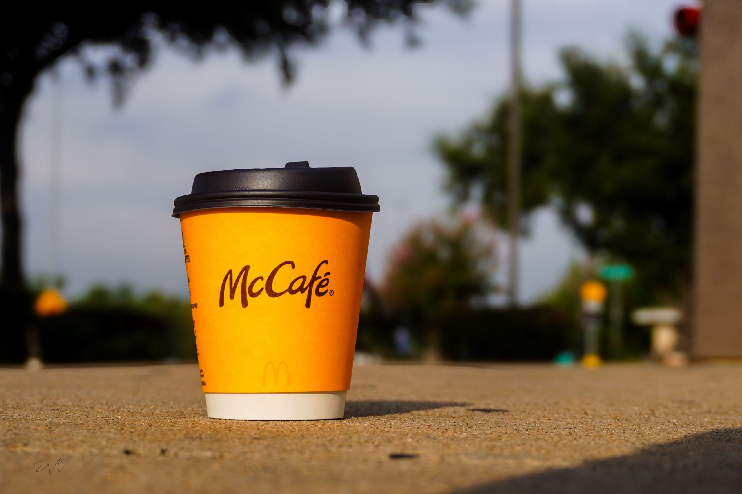 A McCafé coffee cup with a black lid sits on a sunlit sidewalk outdoors, with blurred trees and street signs in the background.