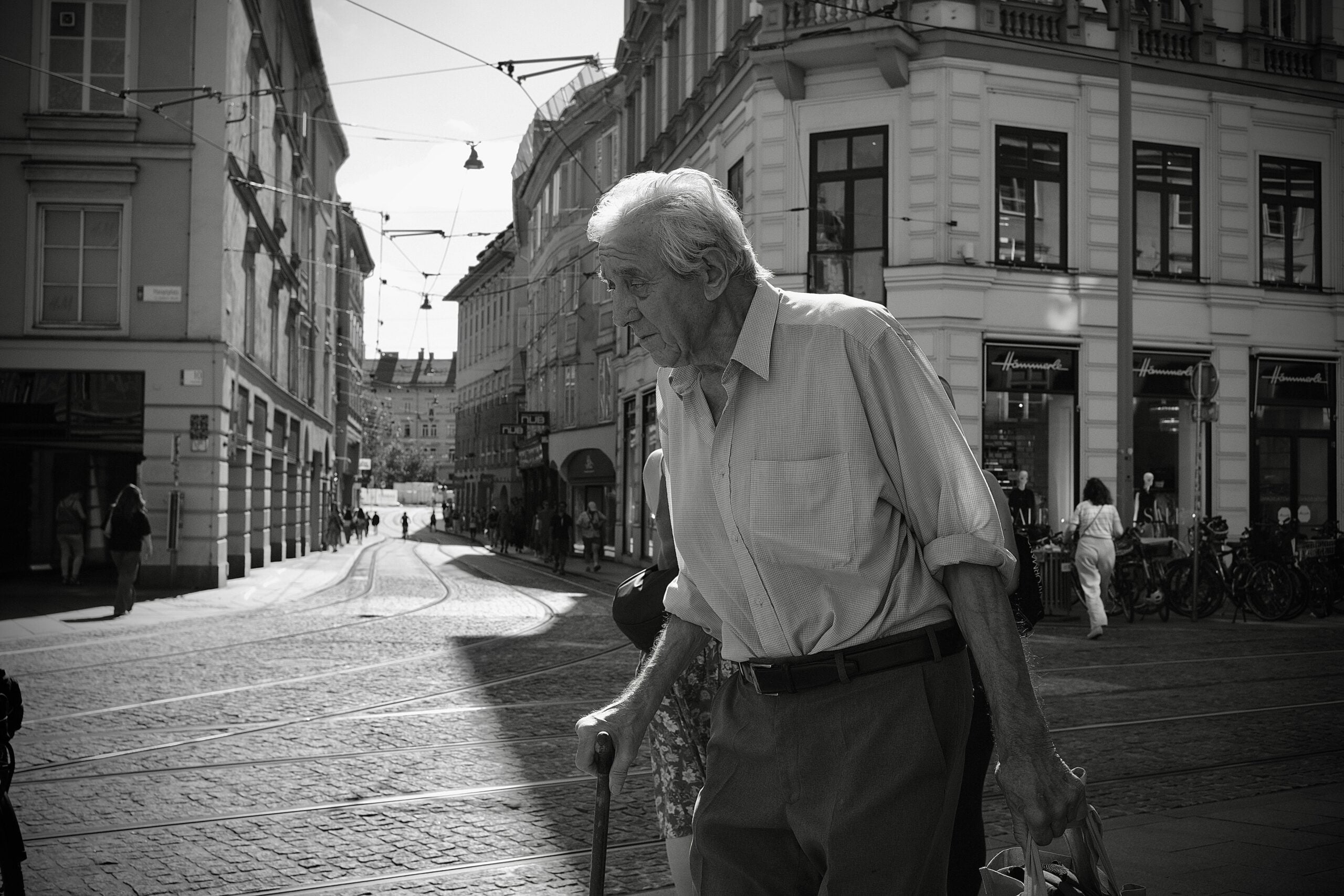 An elderly man with a cane walks across a cobblestone street in a city, surrounded by historic buildings, with sunlight casting shadows and a few people in the background—a quiet moment that could feature in any compilation of Boomer facts highlighting timeless urban traditions.