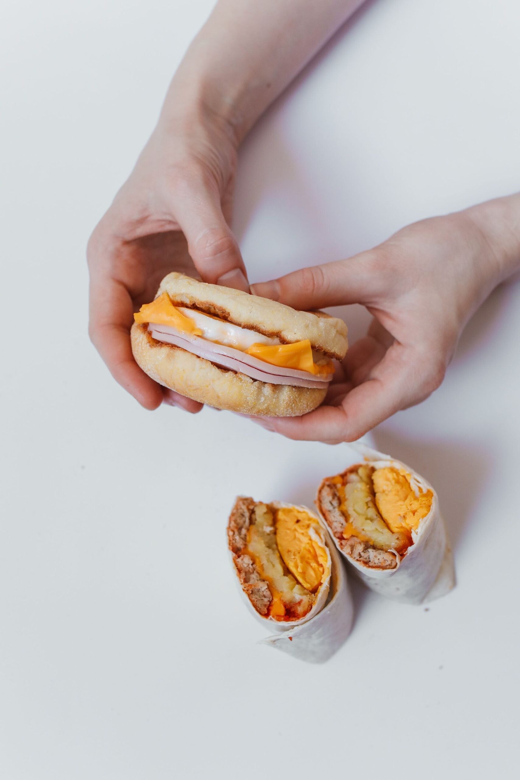 Two hands holding a breakfast sandwich with ham and cheese; below, a halved breakfast burrito with egg, sausage, and cheese on a white surface.