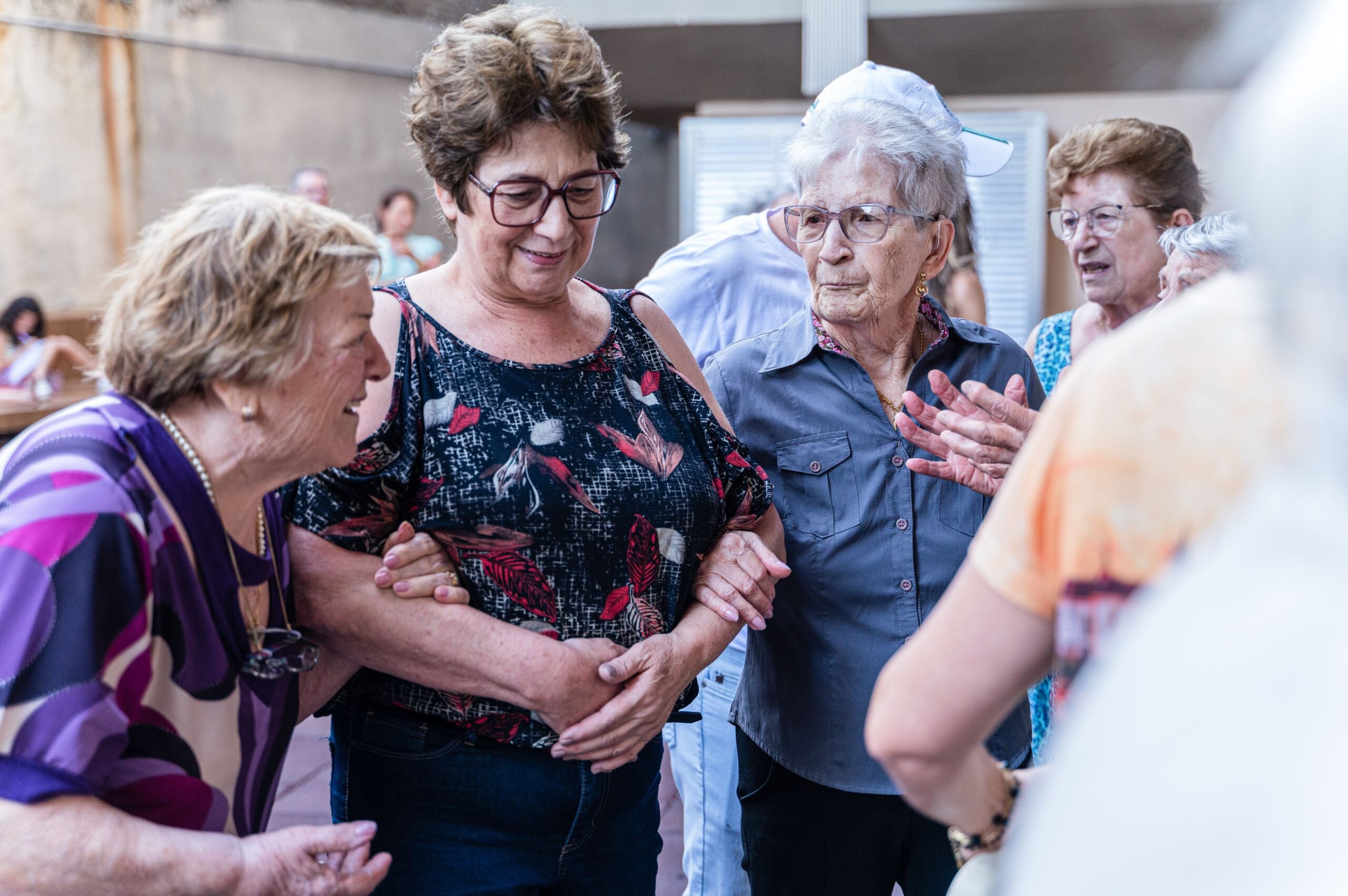 A group of elderly women stand together indoors, smiling and talking as they share some interesting Boomer facts. One woman supports another by holding her arm. The setting appears casual and social.
