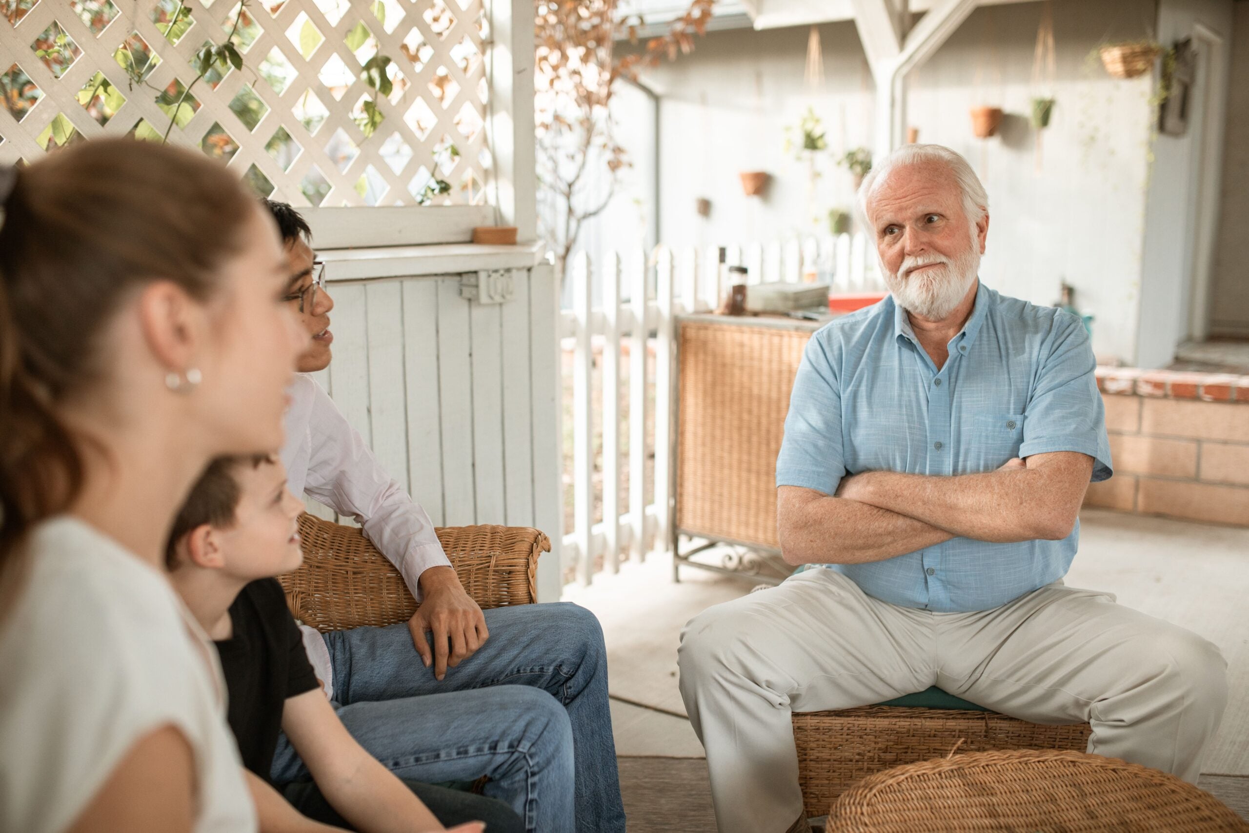An older man with white hair and a beard sits with arms crossed, sharing Boomer facts as he listens to three people, including a child, during a casual conversation on a porch.