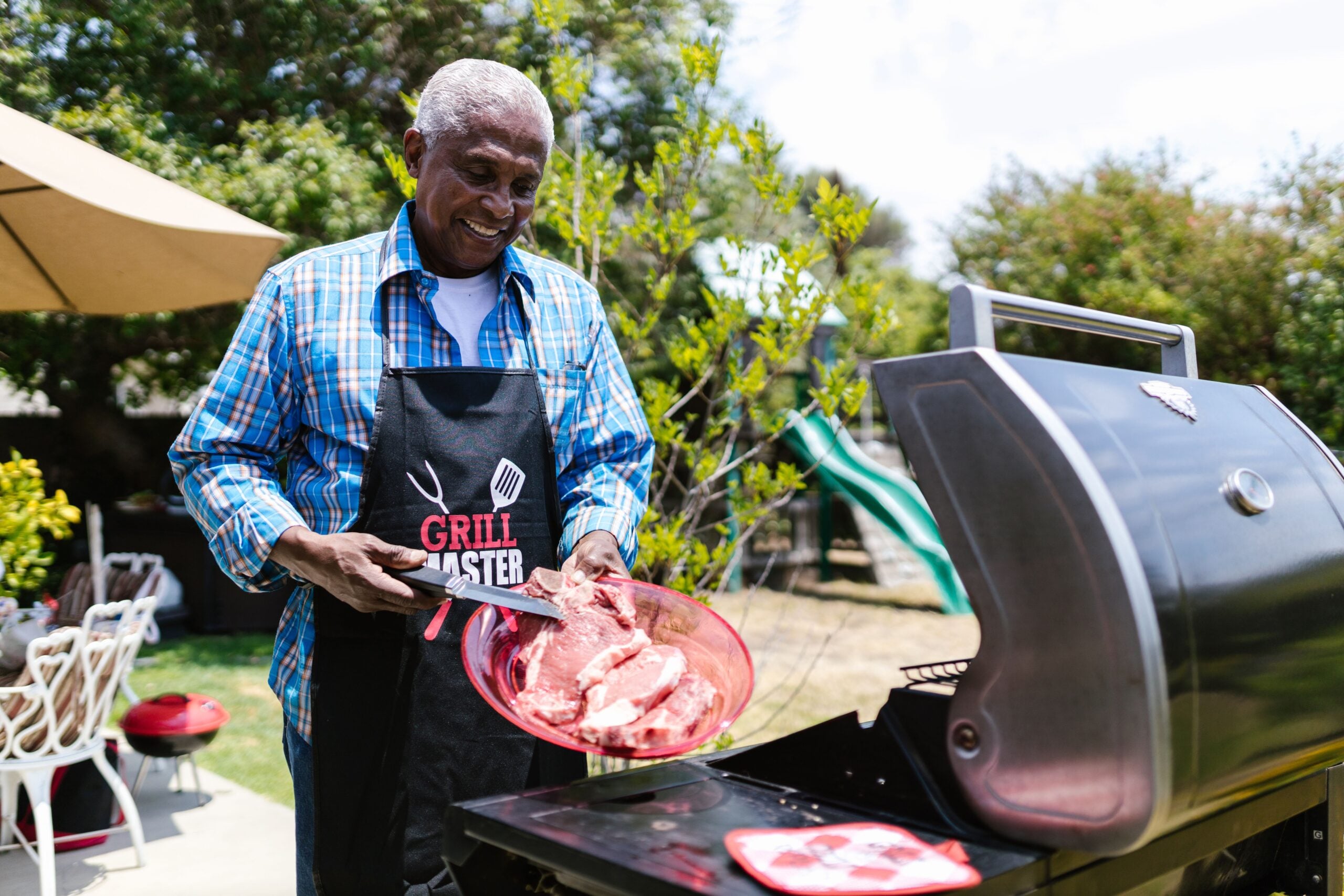 Smiling man in a “Grill Master” apron places raw steaks onto an outdoor barbecue grill in a sunny backyard with patio furniture and greenery in the background, capturing the spirit of Boomers saving money by enjoying home-cooked meals with loved ones.