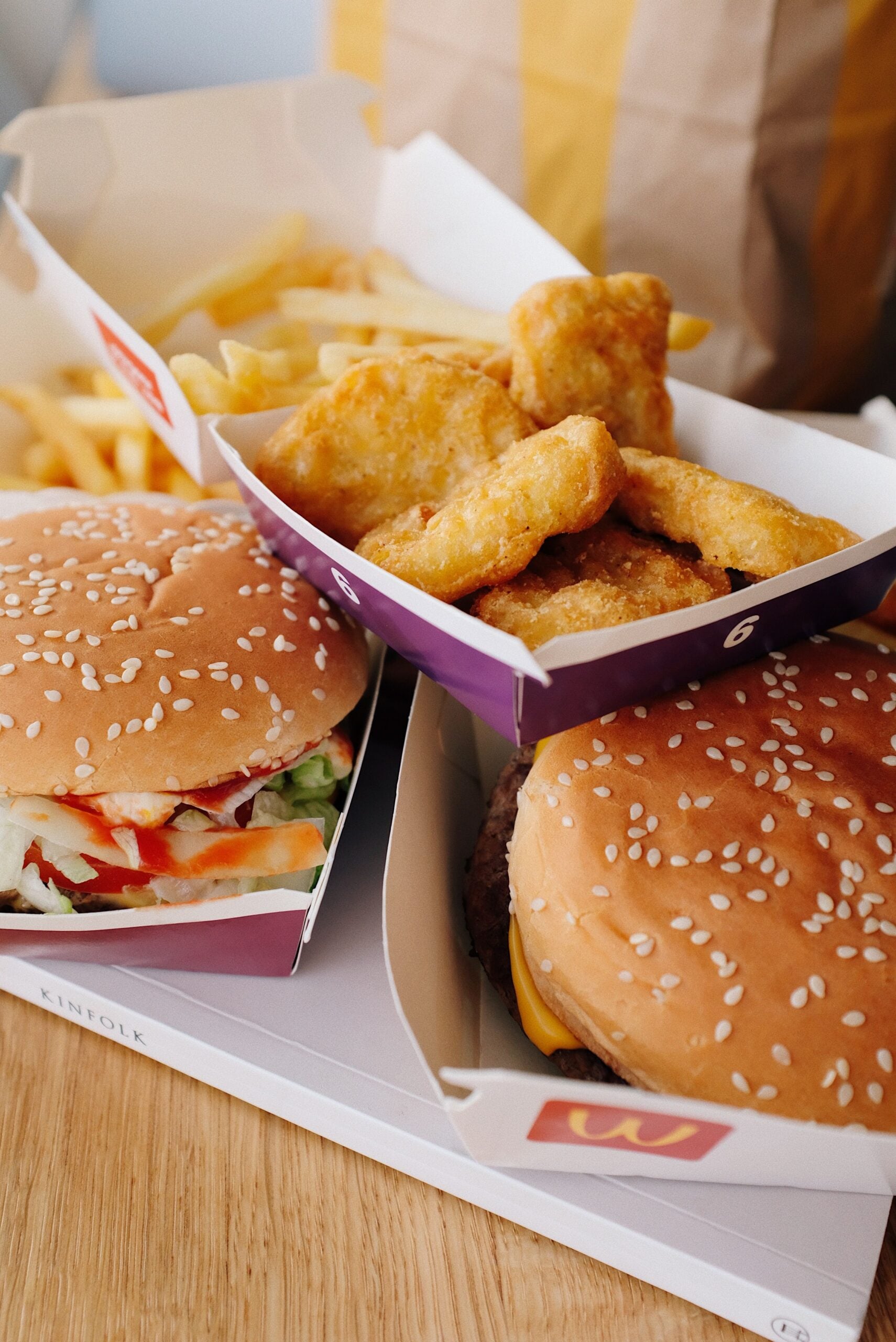 Two burgers with sesame seed buns, crispy chicken nuggets, and a serving of French fries in fast food containers on a wooden surface.