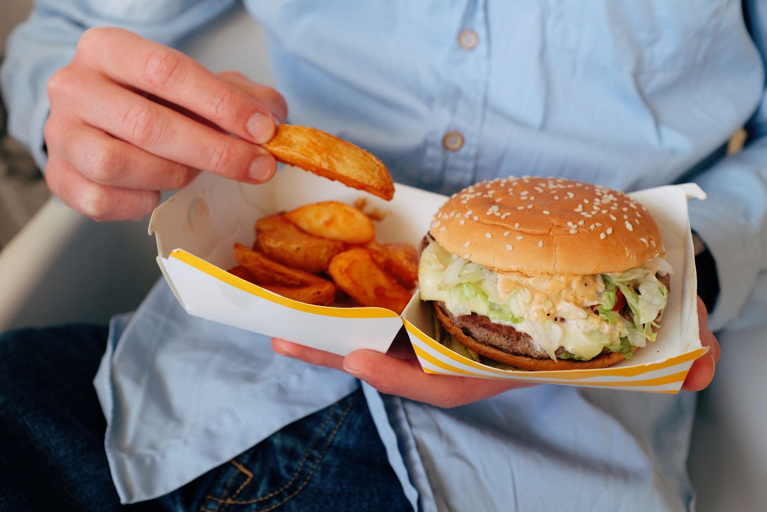 A person in a blue shirt holds a box with a sesame seed hamburger and several potato wedges, picking up one wedge with their hand. Everyday expenses boomers remember.