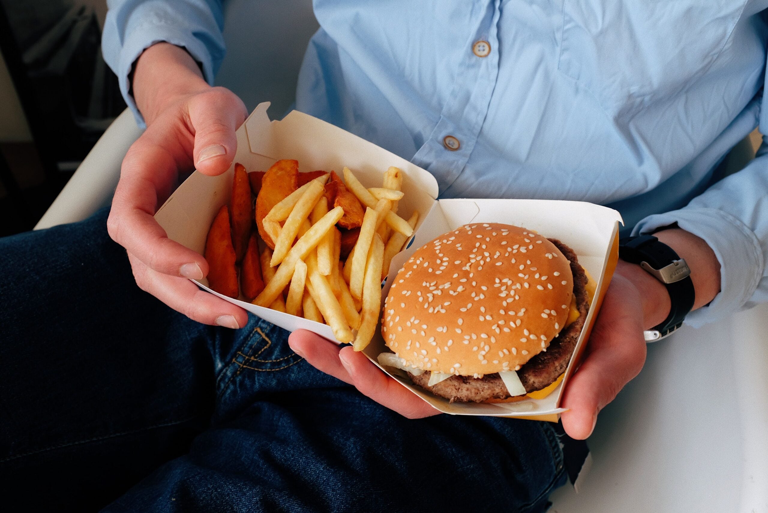 A person in a blue shirt holds an open box with a sesame seed burger, crinkle-cut fries, and potato wedges on their lap.