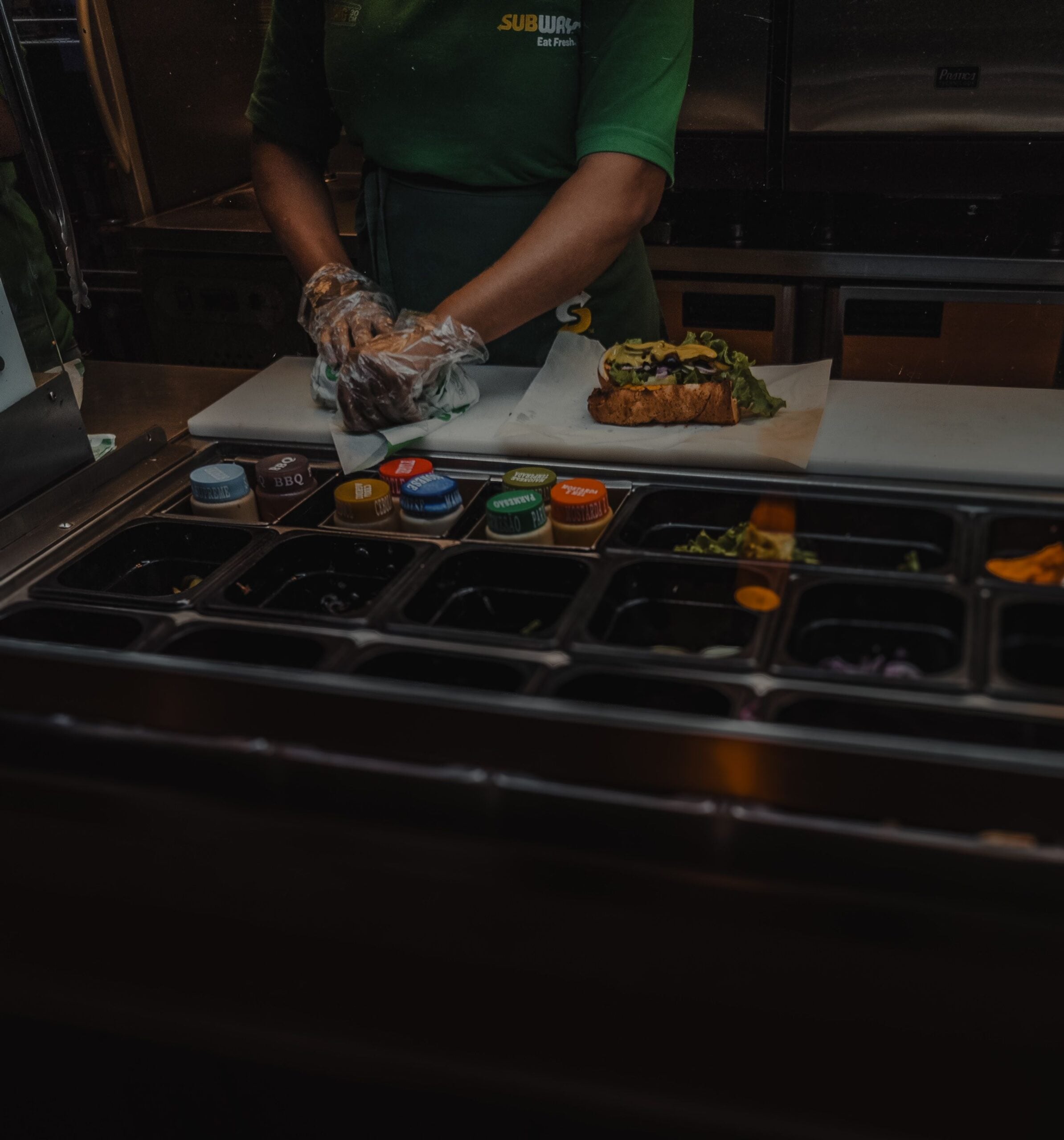 A Subway employee wearing gloves prepares a sandwich behind a counter with various fresh ingredients in containers.