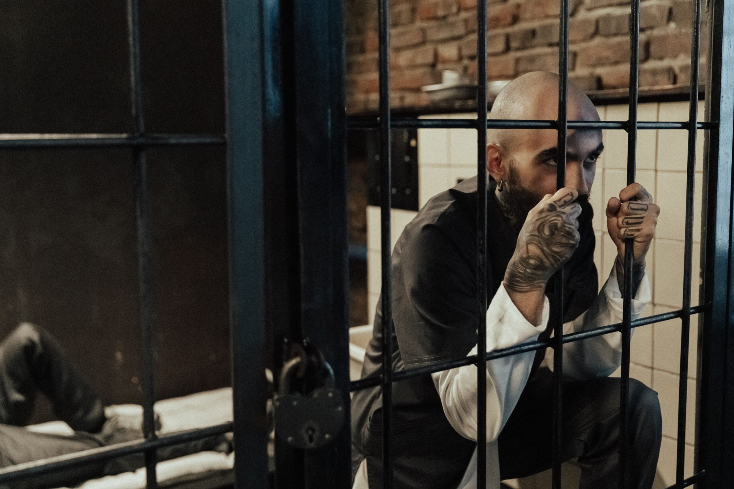 A man with tattoos crouches and grips the bars inside a jail cell, looking outward. Another person lies on a bed in the background. The setting appears tense and somber, capturing the harsh realities of life in 2026 and its unique problems.