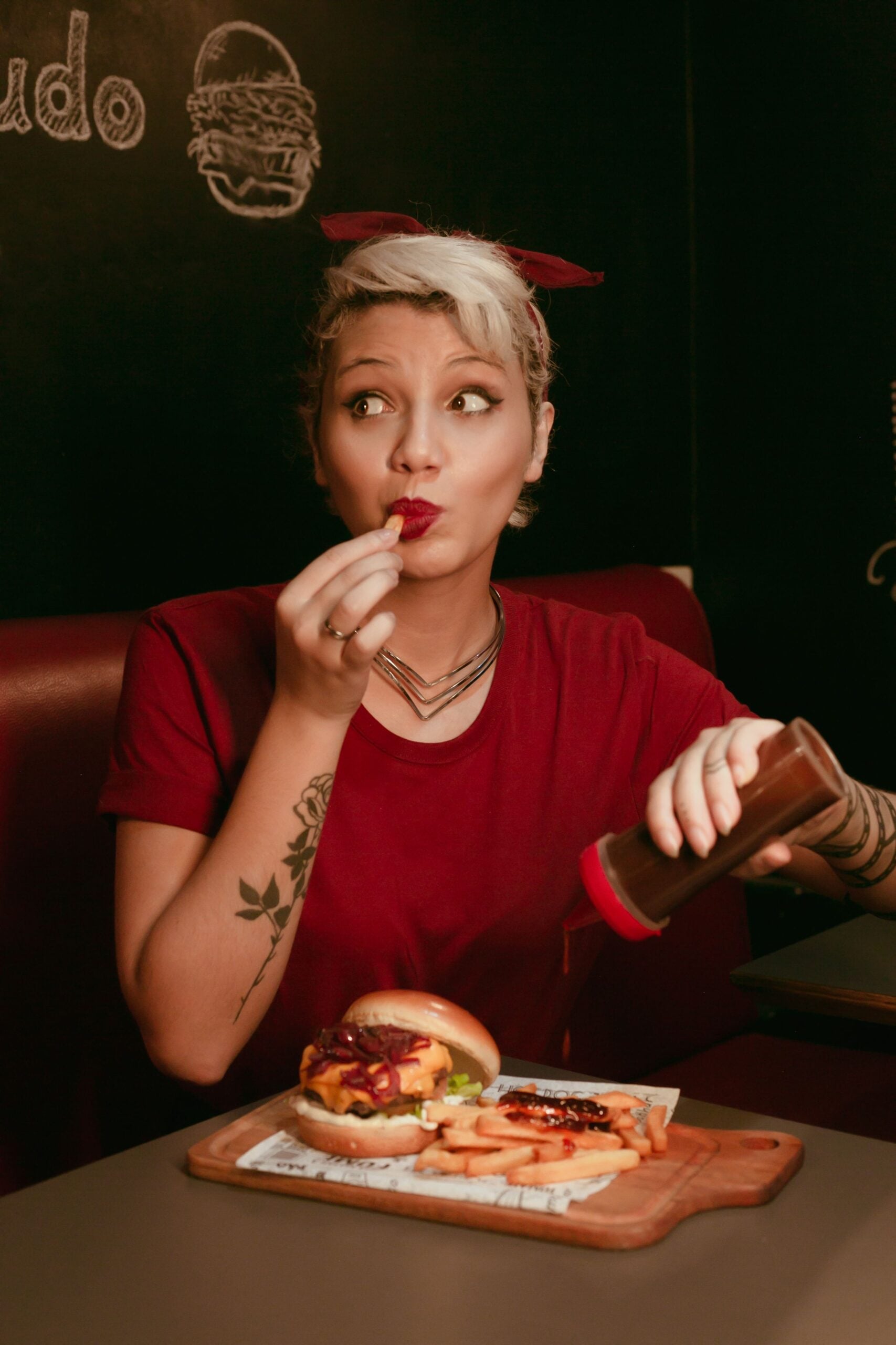 A person in a red shirt puts ketchup on fries, sitting at a table with a burger and fries. They hold a fry near their mouth and look to the side with an amused expression.