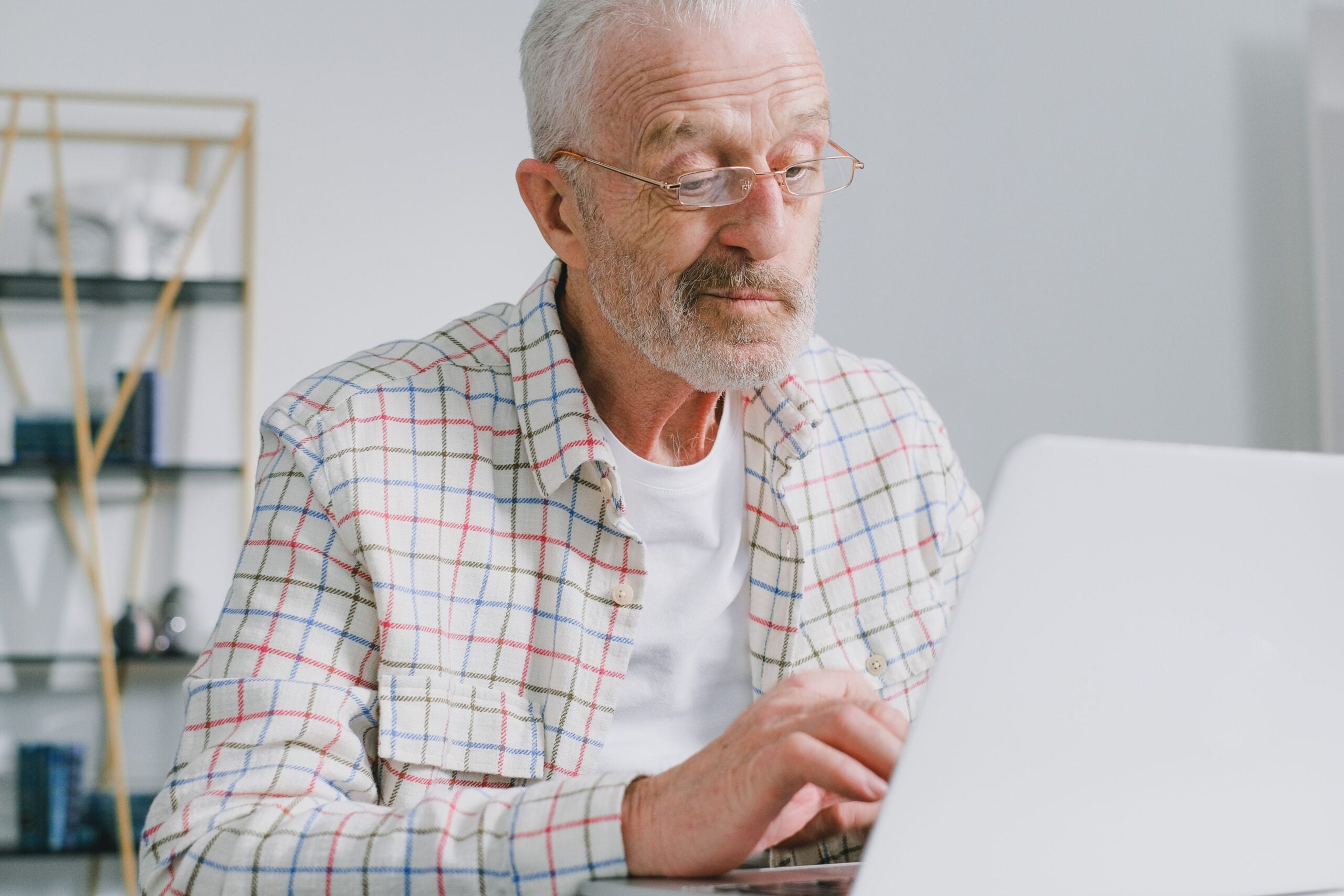 Older man with glasses and a white beard, wearing a plaid shirt, sits at a desk using a laptop in a bright room with shelves in the background, perhaps looking up some intriguing Boomer facts online.