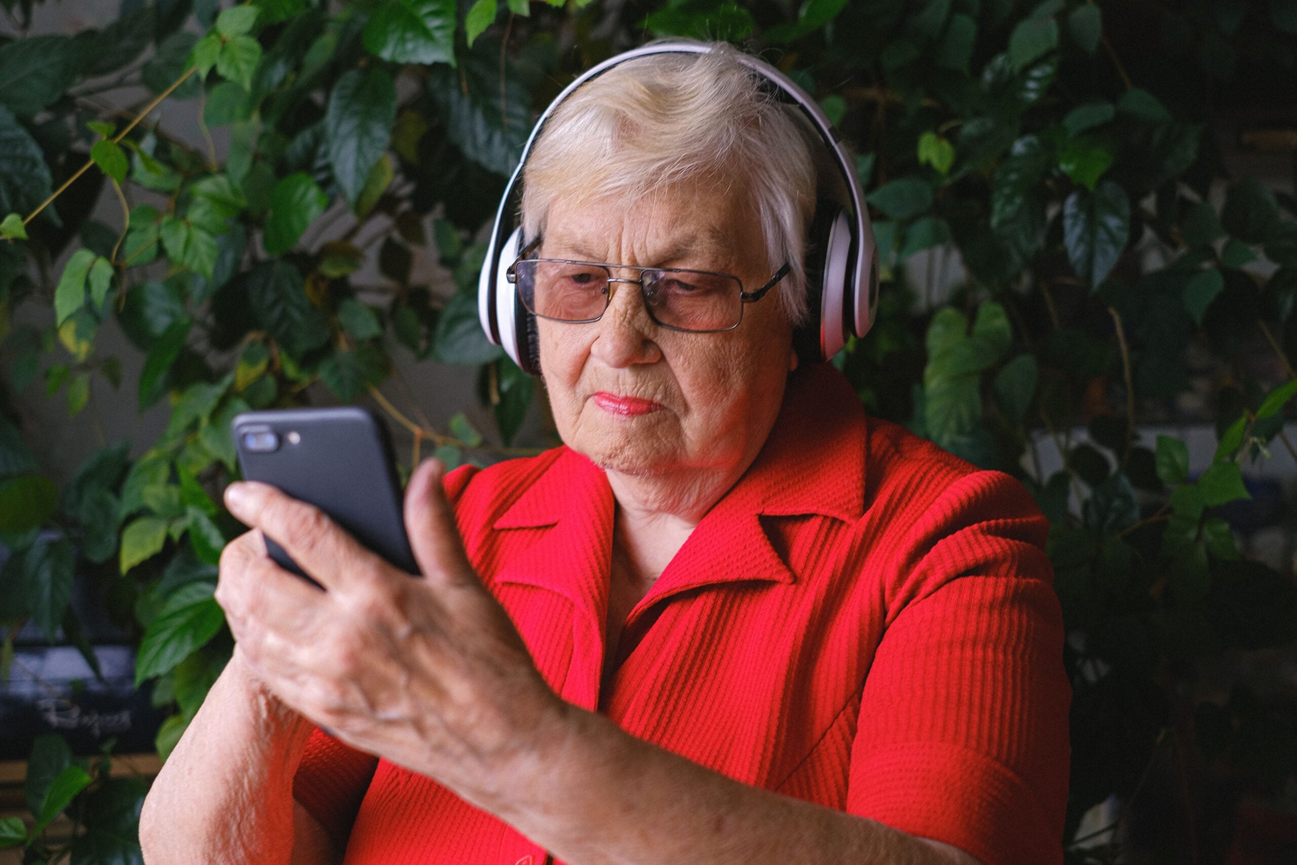 Elderly woman in a red shirt wearing headphones looks at a smartphone, sitting in front of leafy green plants, perhaps discovering some interesting Boomer facts online.