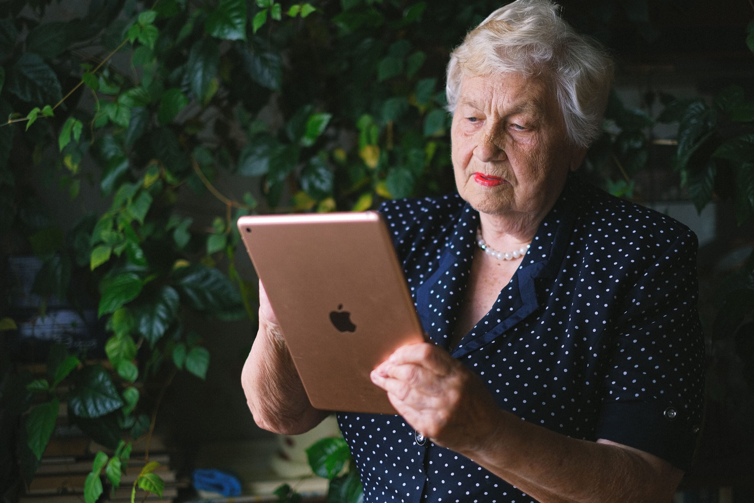 An elderly woman with white hair, wearing a navy blue polka dot blouse, holds and looks at a gold iPad—perhaps reading up on some Boomer facts—against a background of leafy green plants.