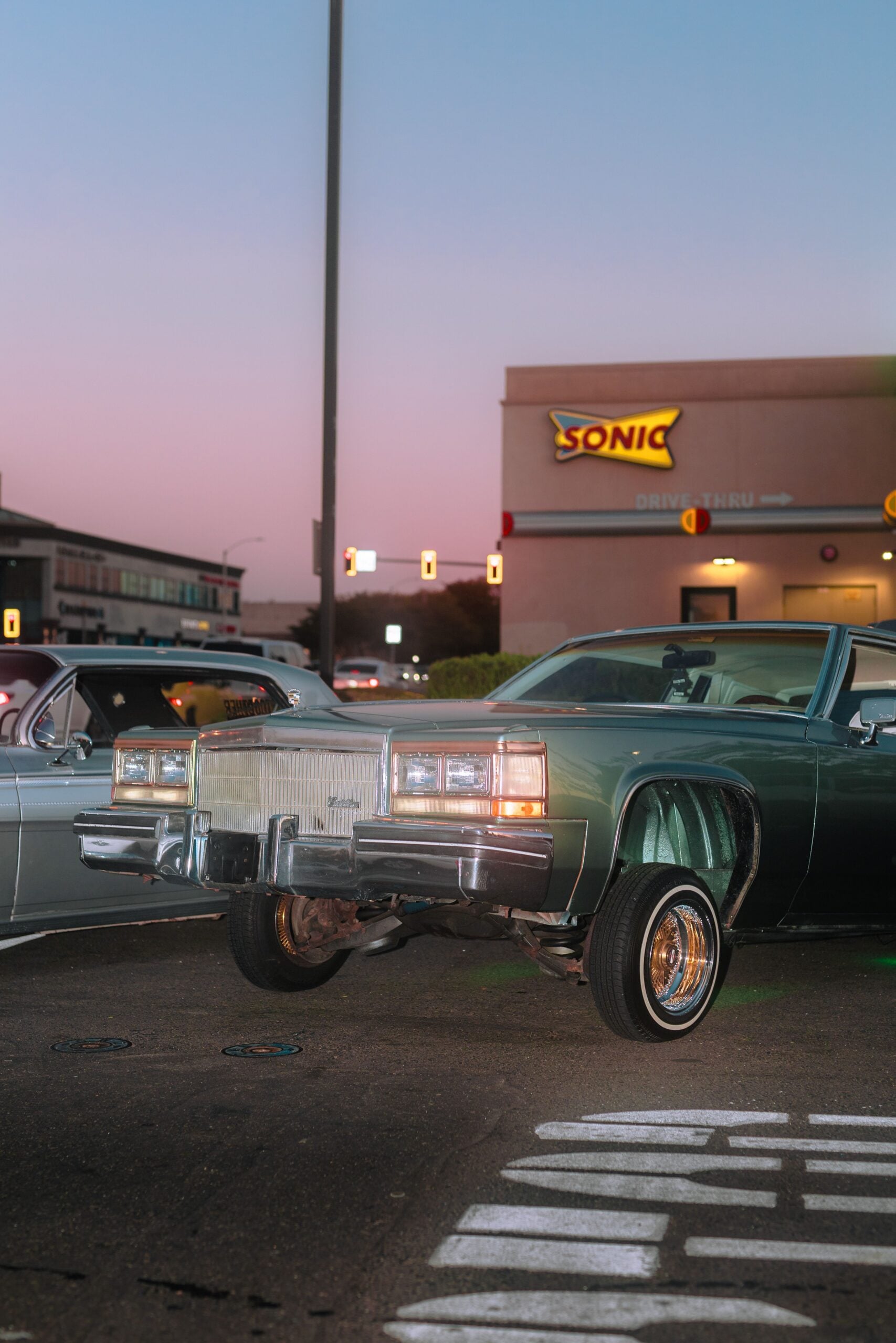 A classic green Cadillac lowrider with its front wheels lifted is parked near a Sonic Drive-In at dusk. Another vintage car is partially visible beside it. The sky has a soft pink gradient.