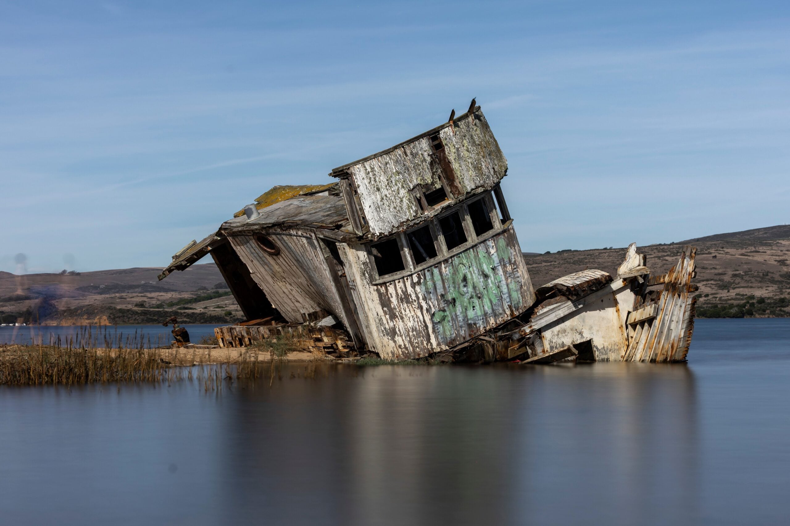 A decaying, graffiti-covered wooden boat rests partially submerged in calm water near a grassy shoreline, with hills and a blue sky in the background—a haunting symbol of life in 2026 problems and the stories left behind.