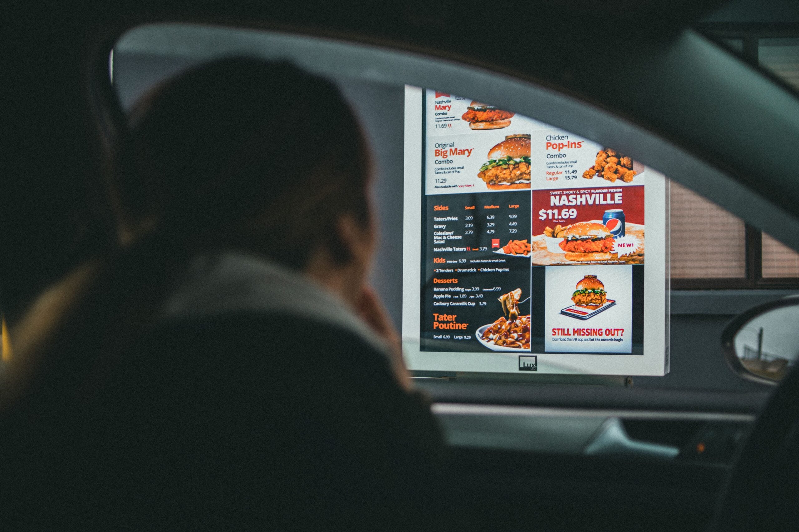 A person in a car looks at a fast-food drive-thru menu displaying chicken meals, prices, and combo options through the driver’s side window.