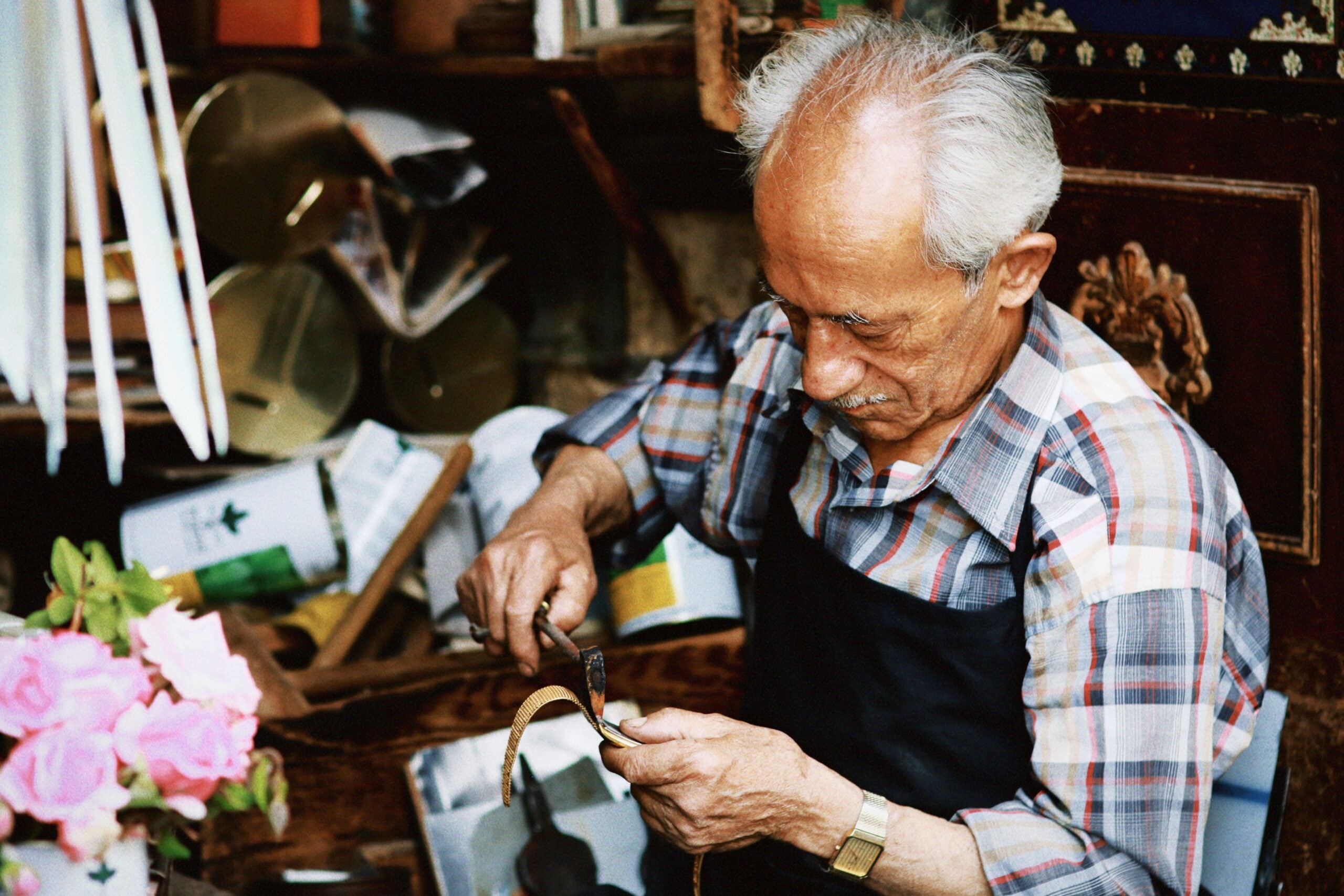 An elderly man in a plaid shirt and apron repairs a leather shoe in a cluttered workshop, surrounded by tools and materials, with pink roses visible in the foreground—a nostalgic scene that brings classic boomer facts to life.