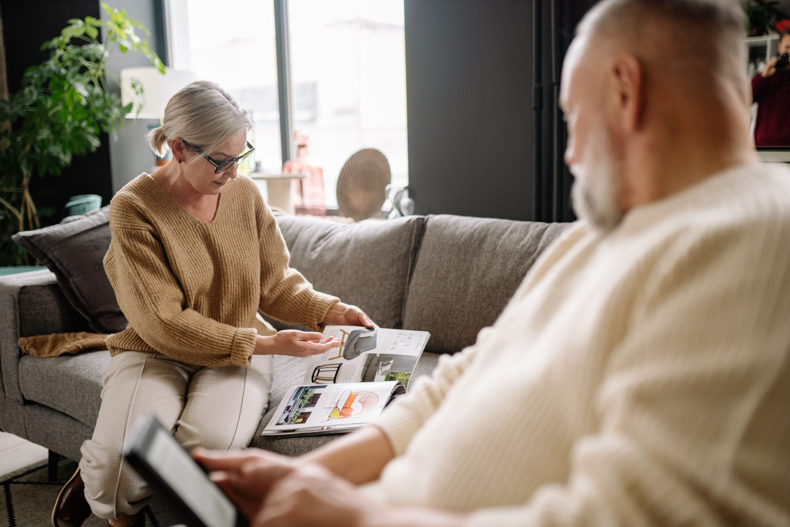 A woman in glasses shows a photo album to a man sitting on a couch. The room is bright with large windows, plants, and cozy decor. As the man holds a tablet, the woman—who enjoys sharing tips about boomers saving money—points to a page in the album, perhaps reminiscing about cherished moments and thrifty habits from years past.