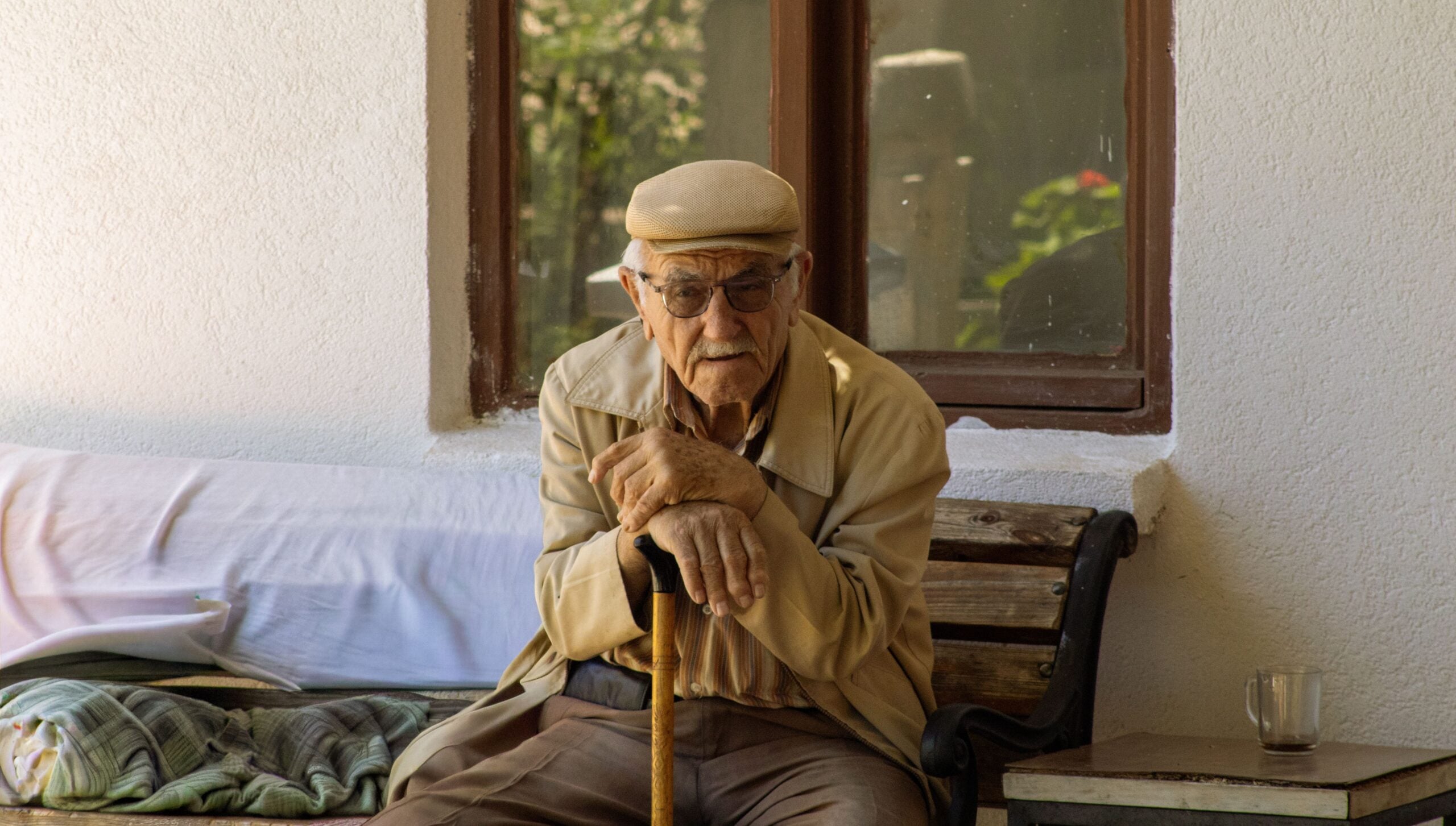 An elderly man wearing a cap and glasses sits on a bench outside, leaning on a cane. As if embodying classic Boomer facts, he is dressed in a beige jacket and brown pants, with a glass mug resting on a small table beside him.