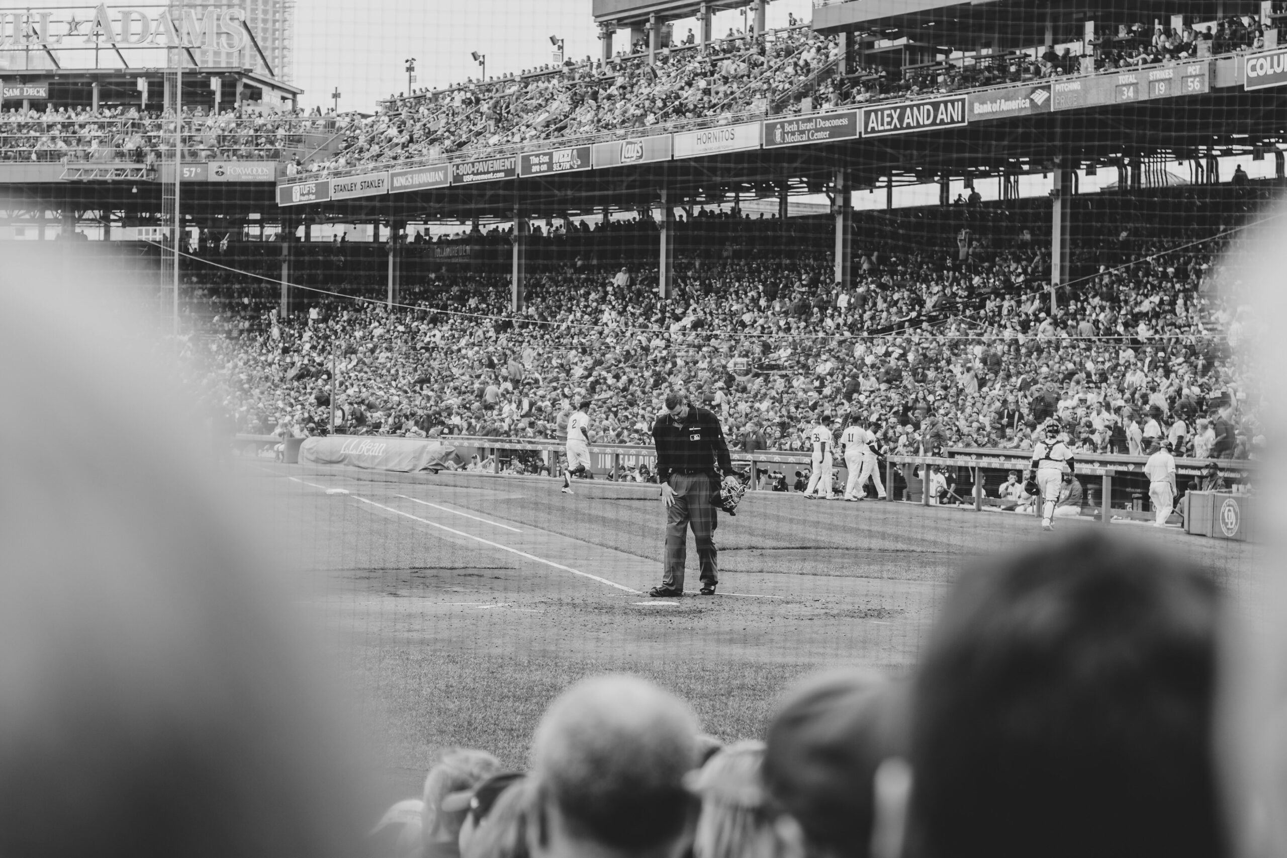 A black and white photo of a baseball game, with an umpire standing near home plate and a large crowd filling the stadium seats in the background. The image is taken from the perspective of the audience.
