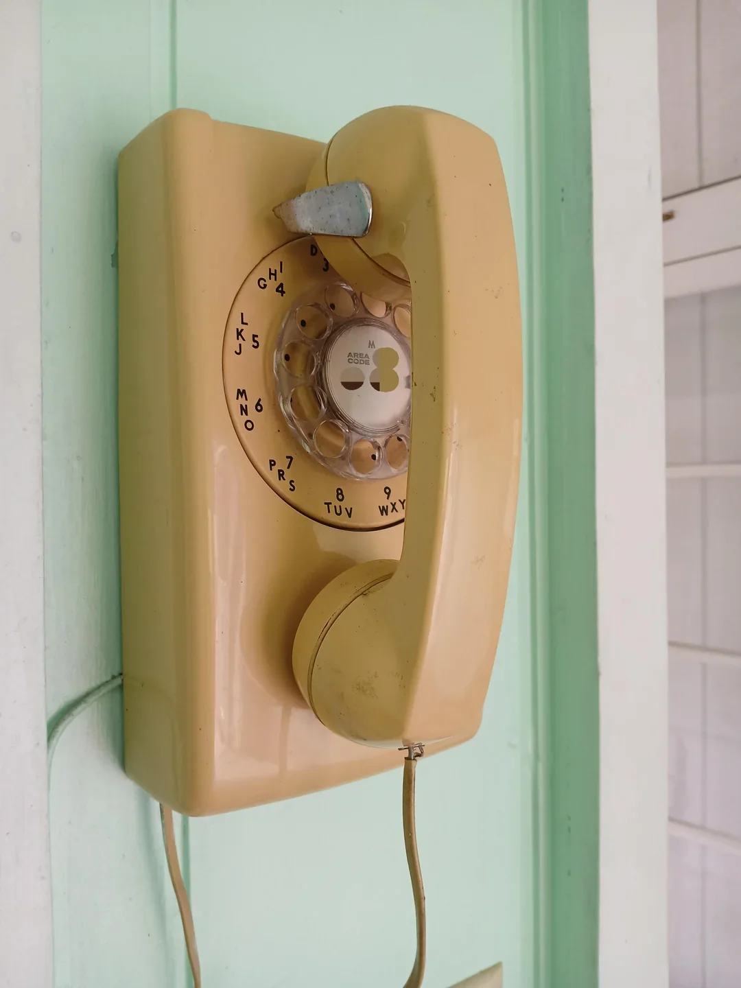 A beige rotary dial wall telephone is mounted on a light green wall. The phone has a corded handset resting on top, and the rotary dial features black numbers and letters.