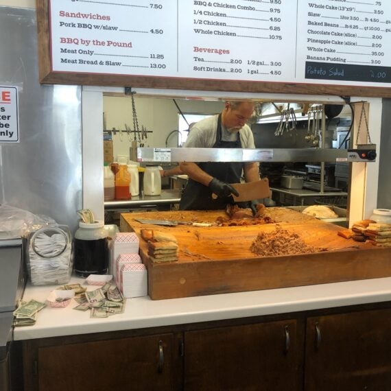 A man slices barbecue meat on a wooden counter in a restaurant kitchen. Slices of bread and piles of cash are on the counter. A menu board hangs above with food and drink options.