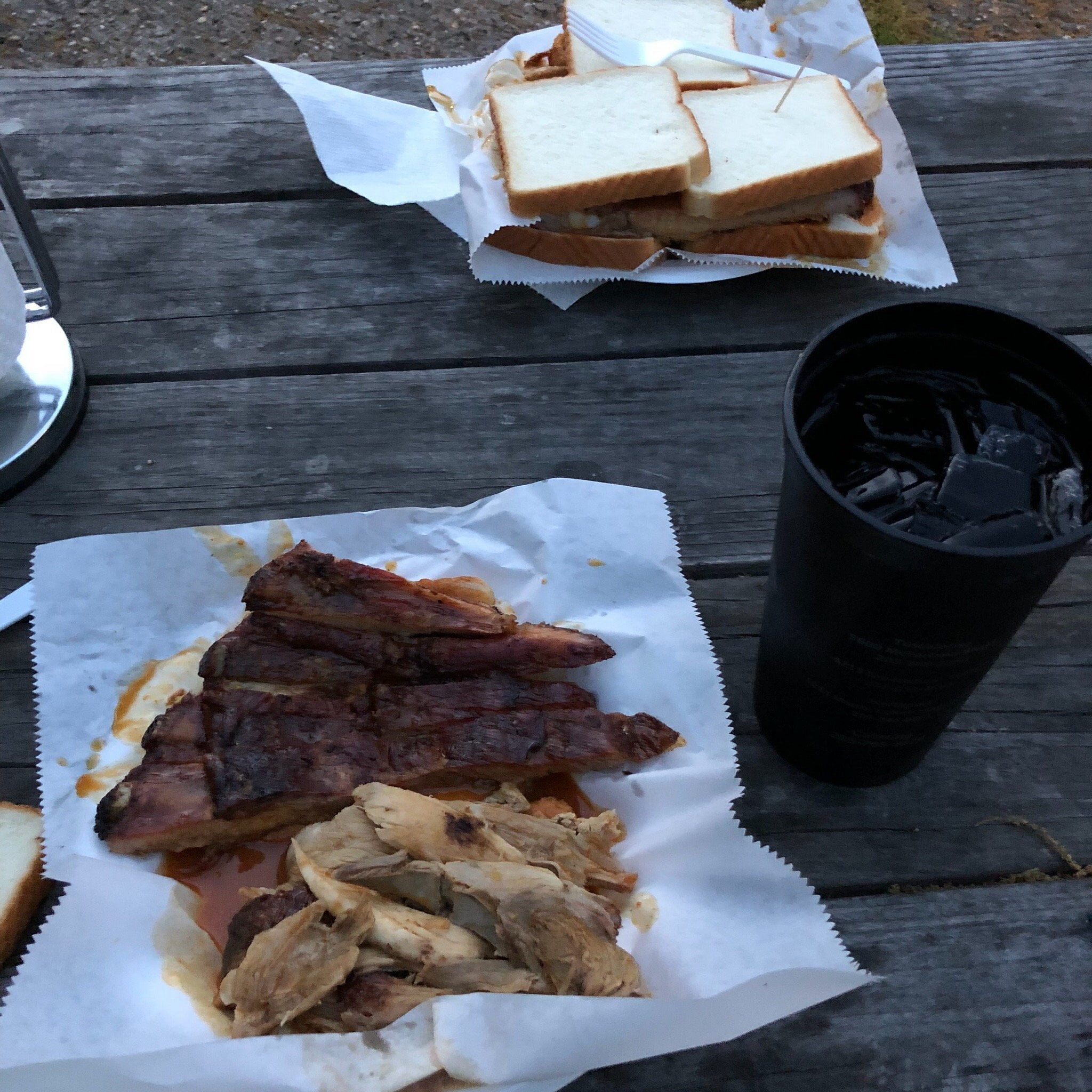 Barbecue meal on a wooden table with ribs, sliced chicken, barbecue sauce, white bread, and a black cup filled with ice.
