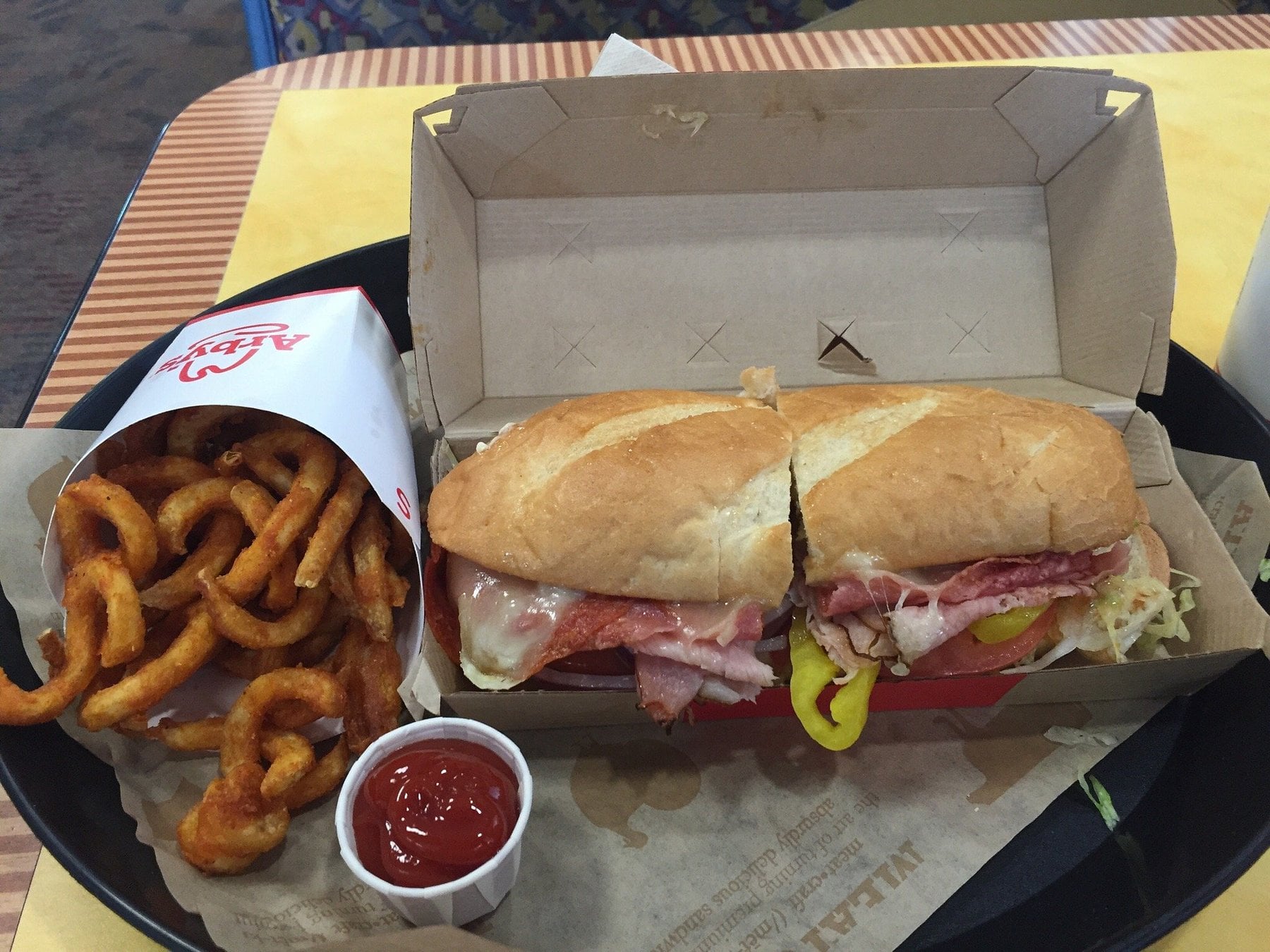 A tray with curly fries in a paper container, a cup of ketchup, and a toasted sub sandwich filled with meats, cheese, and banana peppers in an open box on a yellow table.