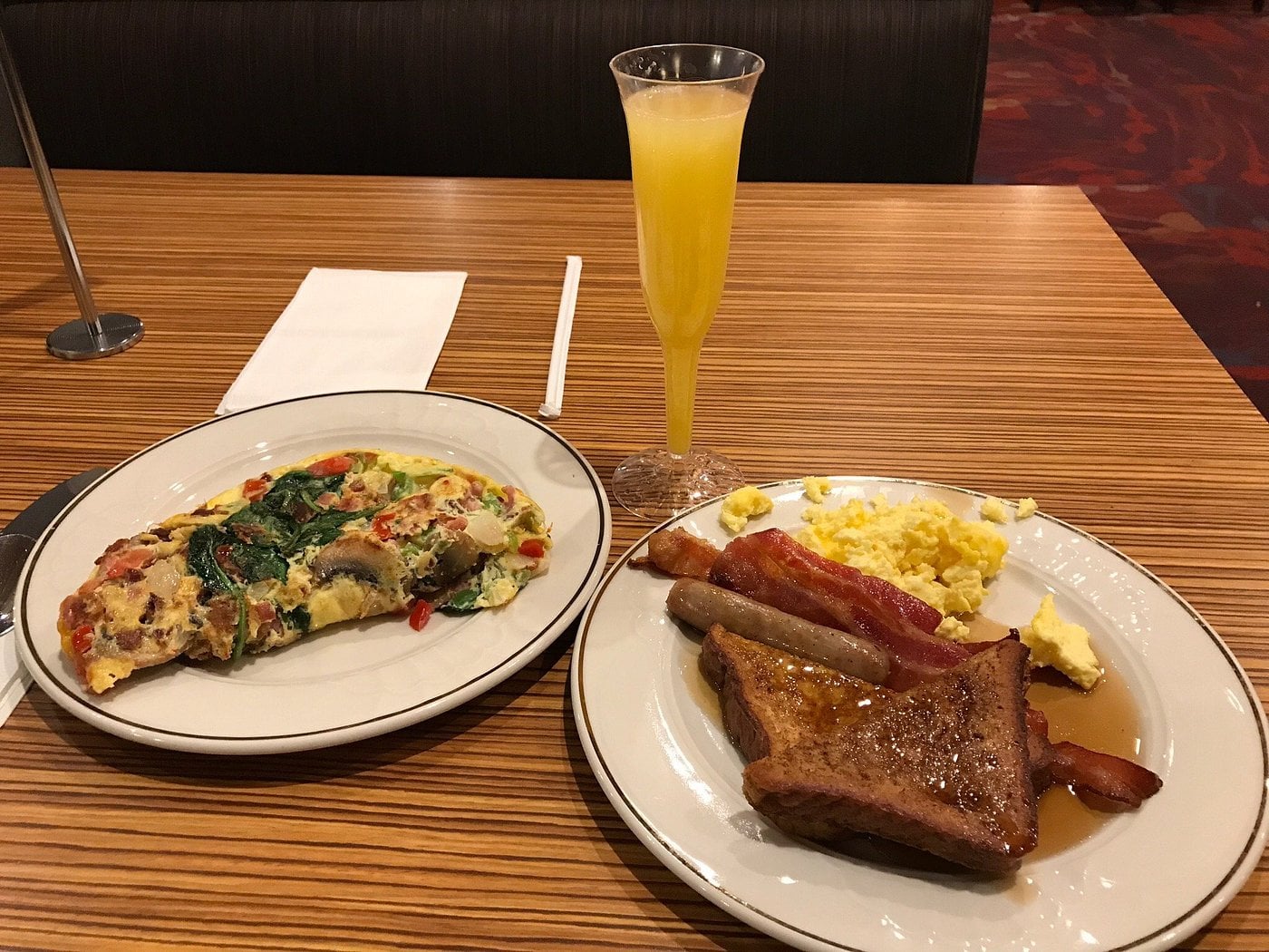 Two plates: one with an omelet, the other with scrambled eggs, bacon, sausage, and French toast. A glass of orange juice is behind the plates on a wooden table.
