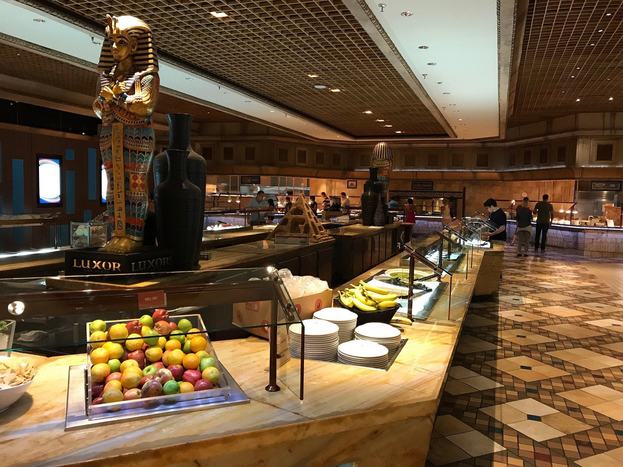 A buffet with fruit, plates, and Egyptian-themed statues, including one of a pharaoh, in a spacious, well-lit dining area with a patterned tile floor.