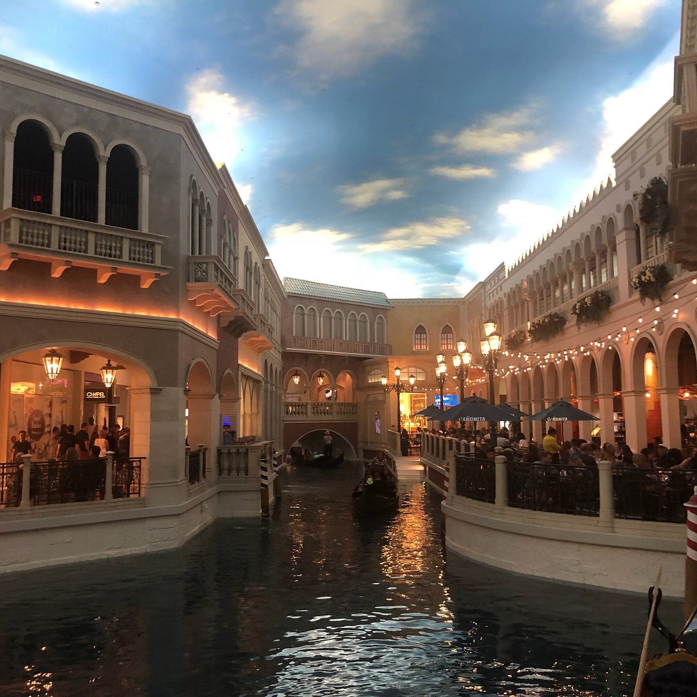 Indoor Venetian canal scene with gondola, elegant buildings, arched bridges, and people dining beside the water under a painted sky ceiling.