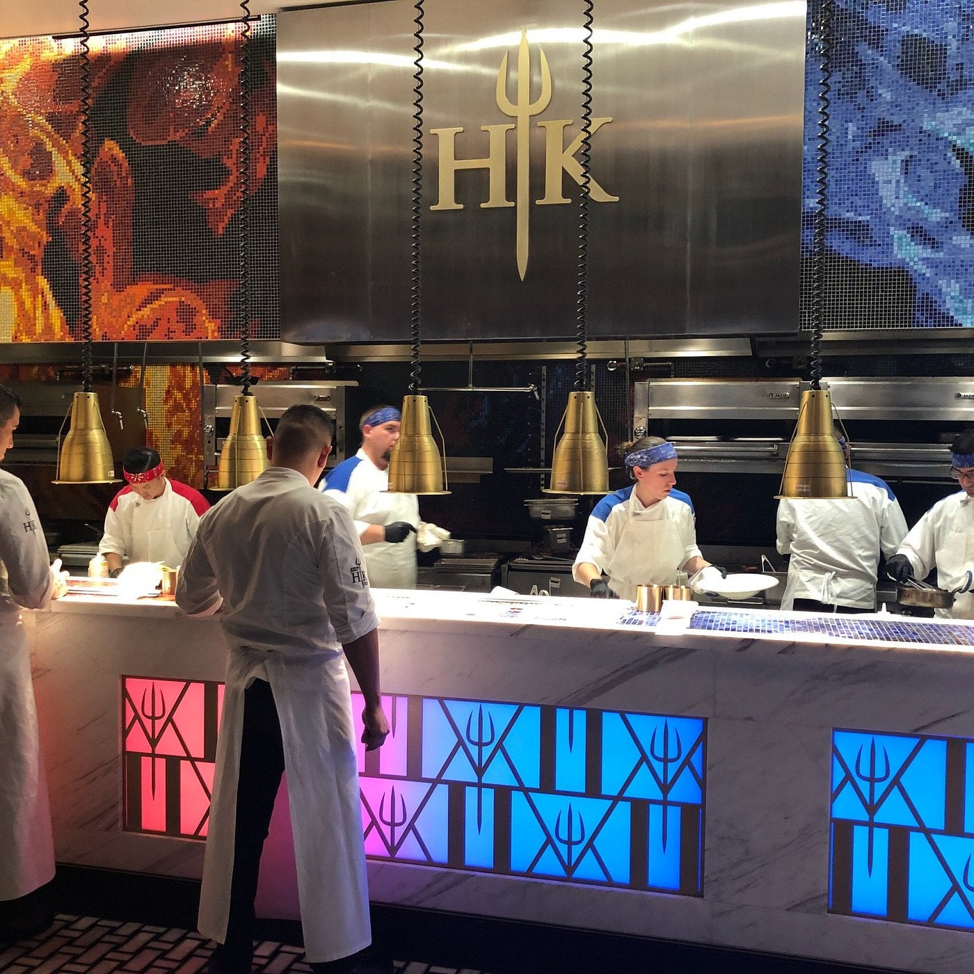Chefs in white uniforms work in a restaurant kitchen with “HK” and a trident logo on the wall. The counter glows with blue and red lights, matching decorations behind the chefs.