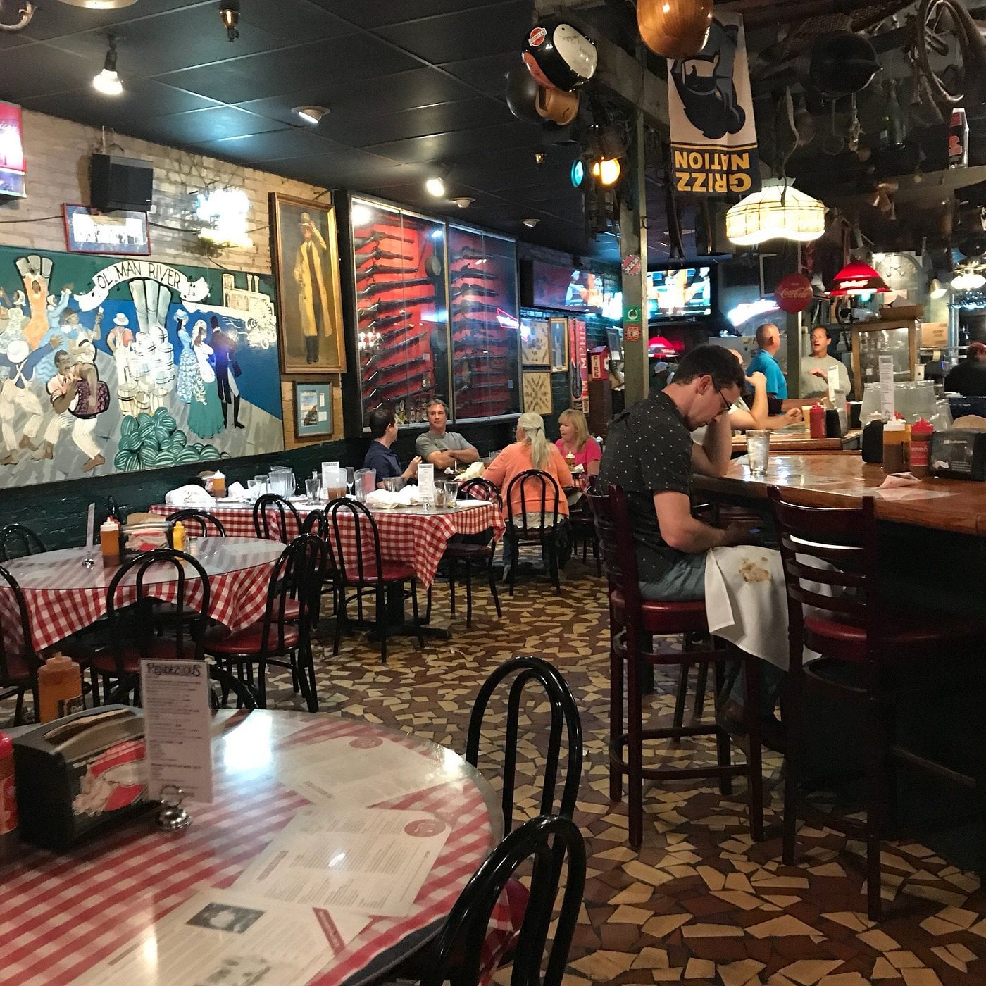 A cozy restaurant with checkered tablecloths, wall art, and sports decor. Several people are dining, and a man sits alone at the bar. The floor has a mosaic pattern, and the atmosphere appears casual and lively.