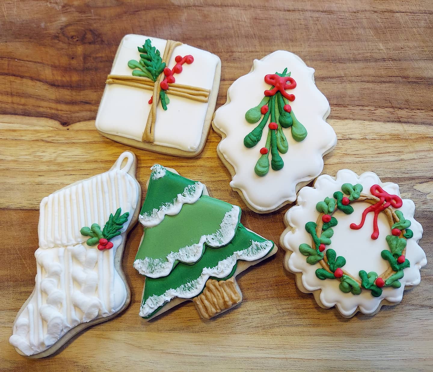 Five Christmas-themed decorated sugar cookies on a wooden surface: a stocking, a gift, a tree, a plaque with mistletoe, and a wreath with a red bow and holly berries—perfect examples of delightful homemade Christmas treats.